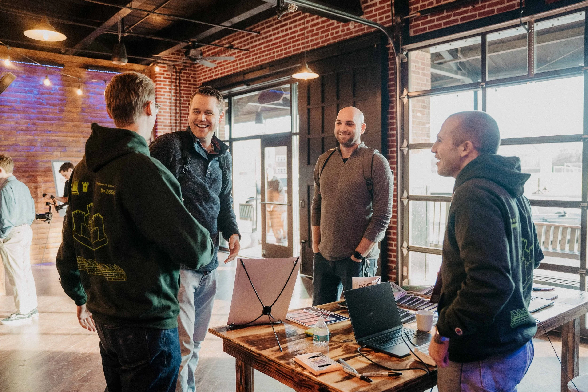 Four men are standing and talking inside a modern industrial-style building with brick walls, large windows, and warm lighting. Two of them are wearing black hoodies with yellow graphics, and the other two are dressed casually in jackets and hoodies.