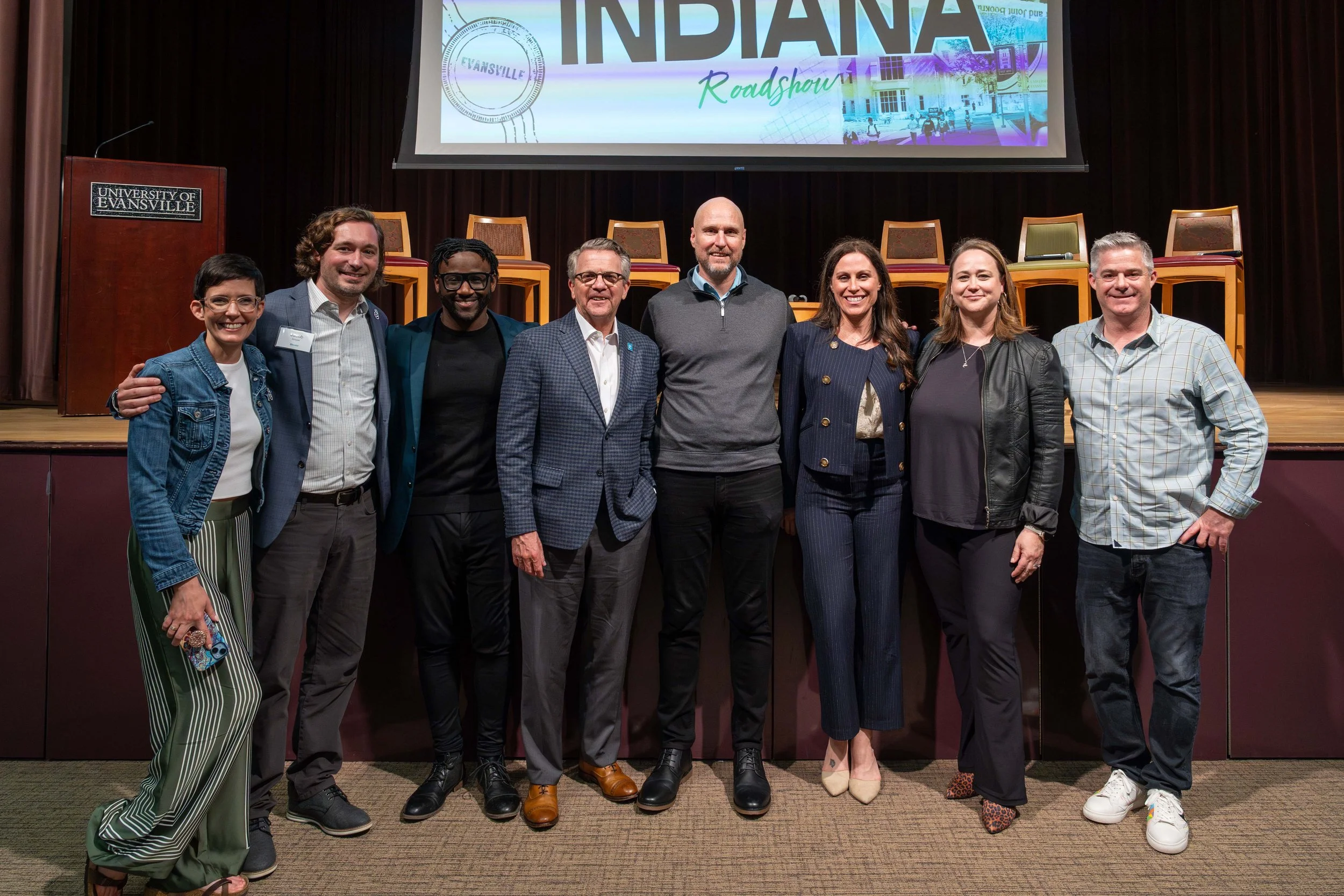 Group of nine diverse individuals standing together on stage, smiling for a photo, in front of a large screen displaying 'Indiana Roadshow' and a university podium with a logo that reads 'University of Evansville'