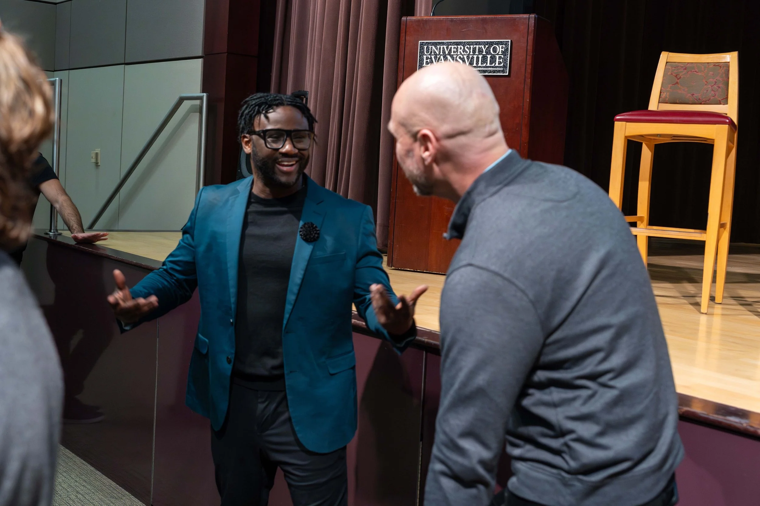 Two men engaged in conversation in an auditorium with a sign that reads 'University of Evansville' on stage, one wearing a blue blazer and glasses, the other in a gray shirt.
