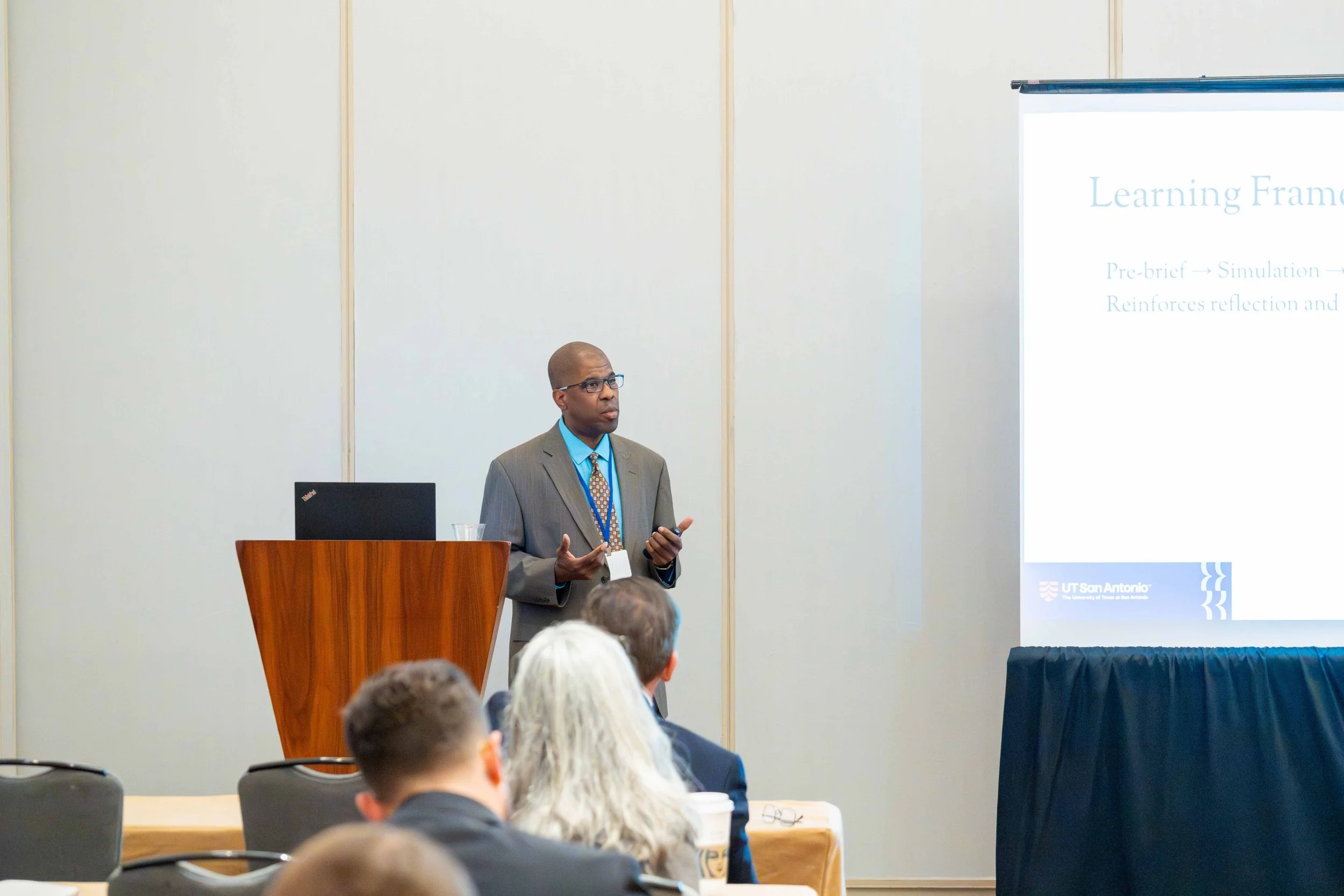 A man in a gray suit, blue shirt, and orange tie giving a presentation at a conference, standing behind a wooden podium with a laptop, with an audience seated in front. The presentation slide on the screen reads 'Learning Frame' and mentions simulati