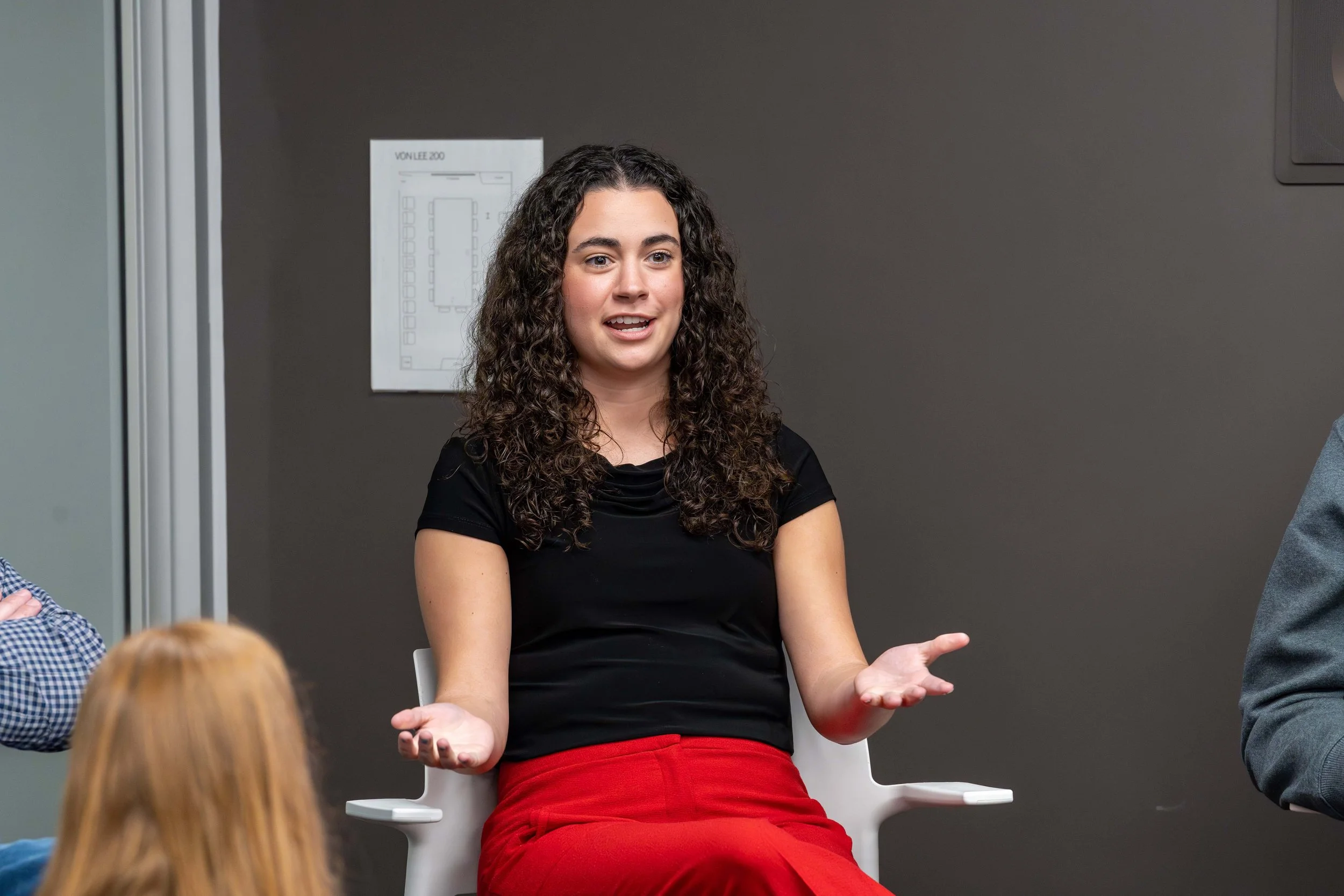 Woman with curly hair wearing a black top and red pants speaking in a discussion or presentation, seated on a white chair in a modern office setting.