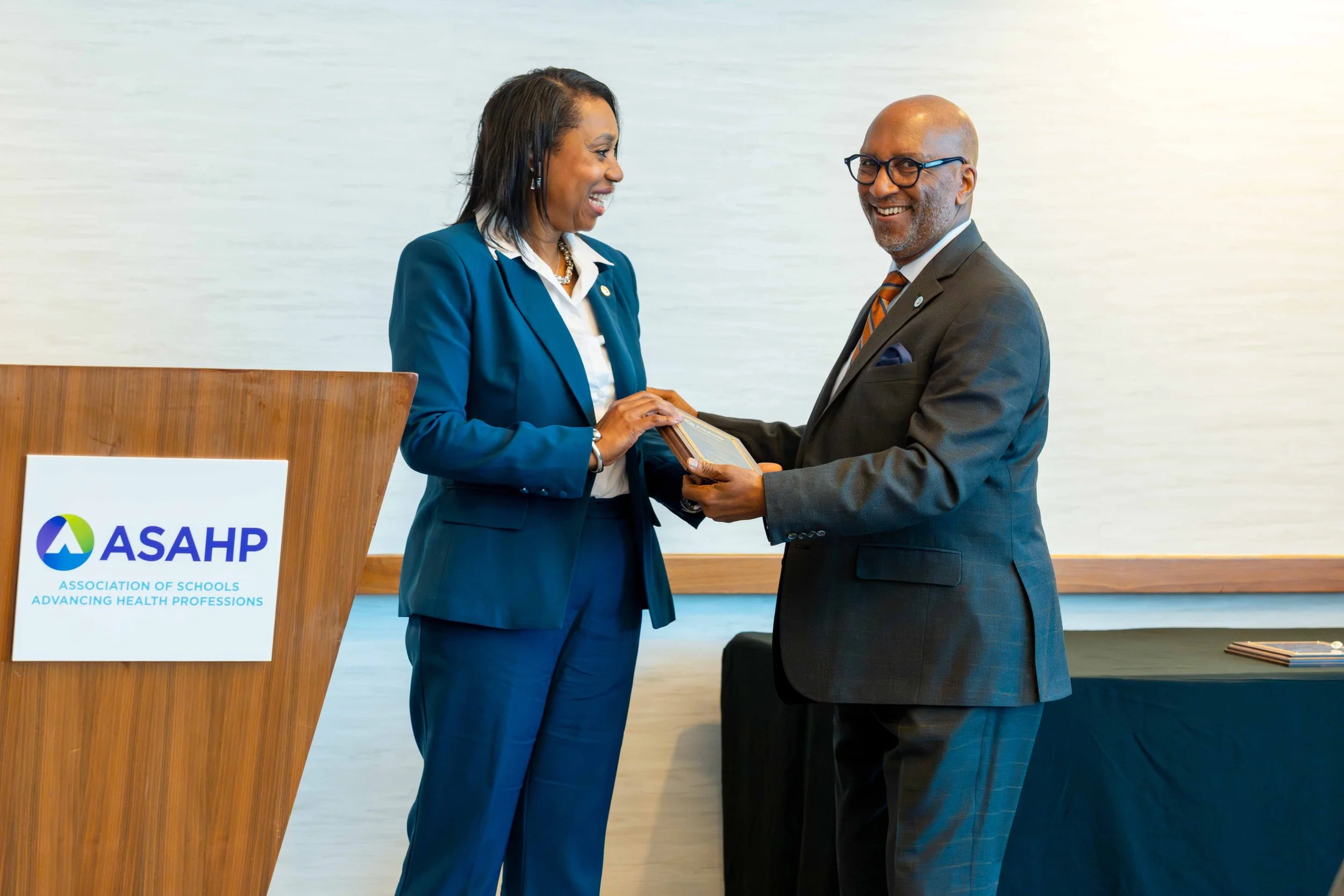 A woman in a blue suit and a man in a dark gray suit exchanging a plaque or award at a conference. There is a podium with a sign that reads 'ASAH' and 'Association of Schools Advancing Health Professions' in the foreground.