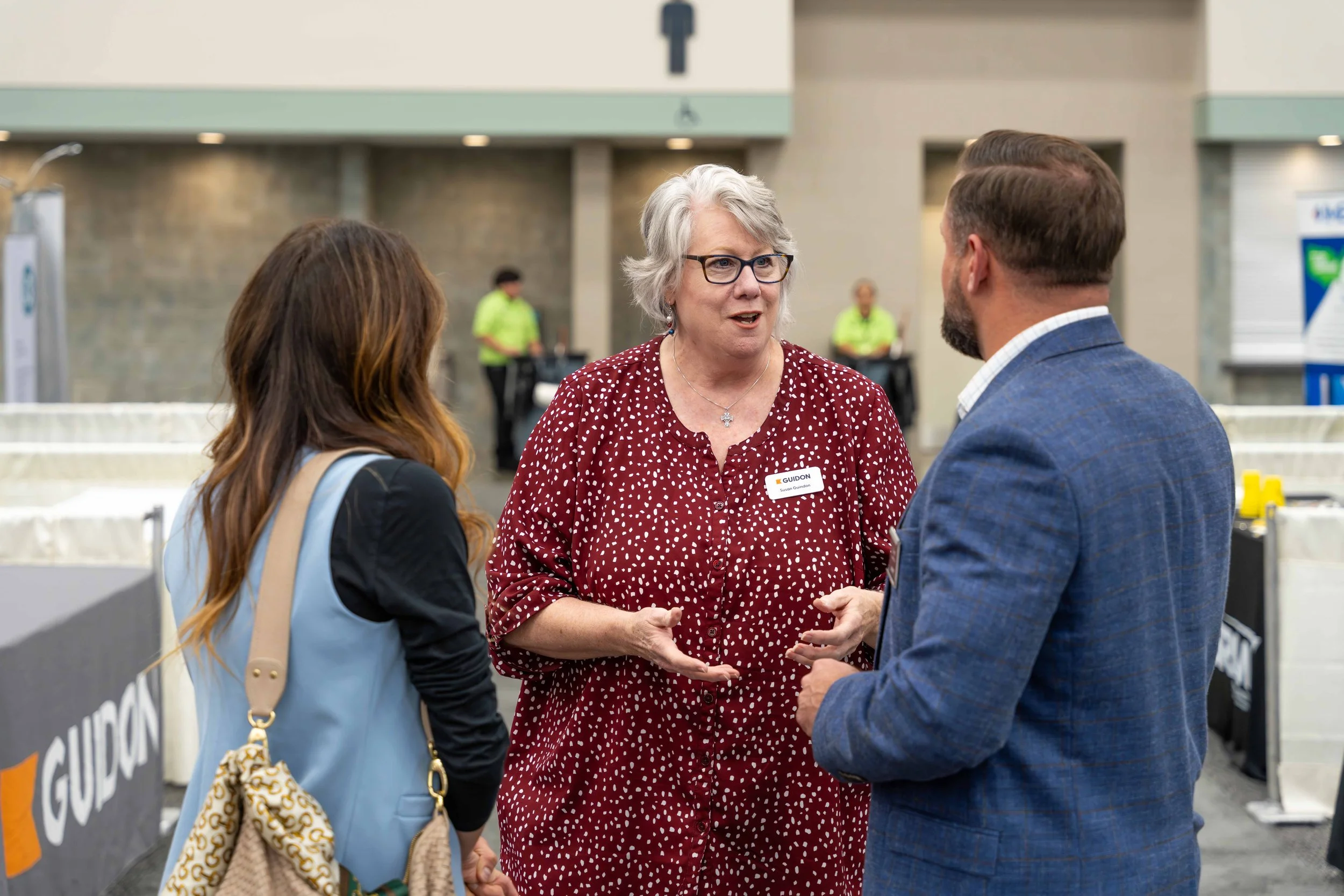 A woman with gray hair and glasses talking to a man and a woman at an event in a large indoor space with signs, security personnel, and booths in the background.
