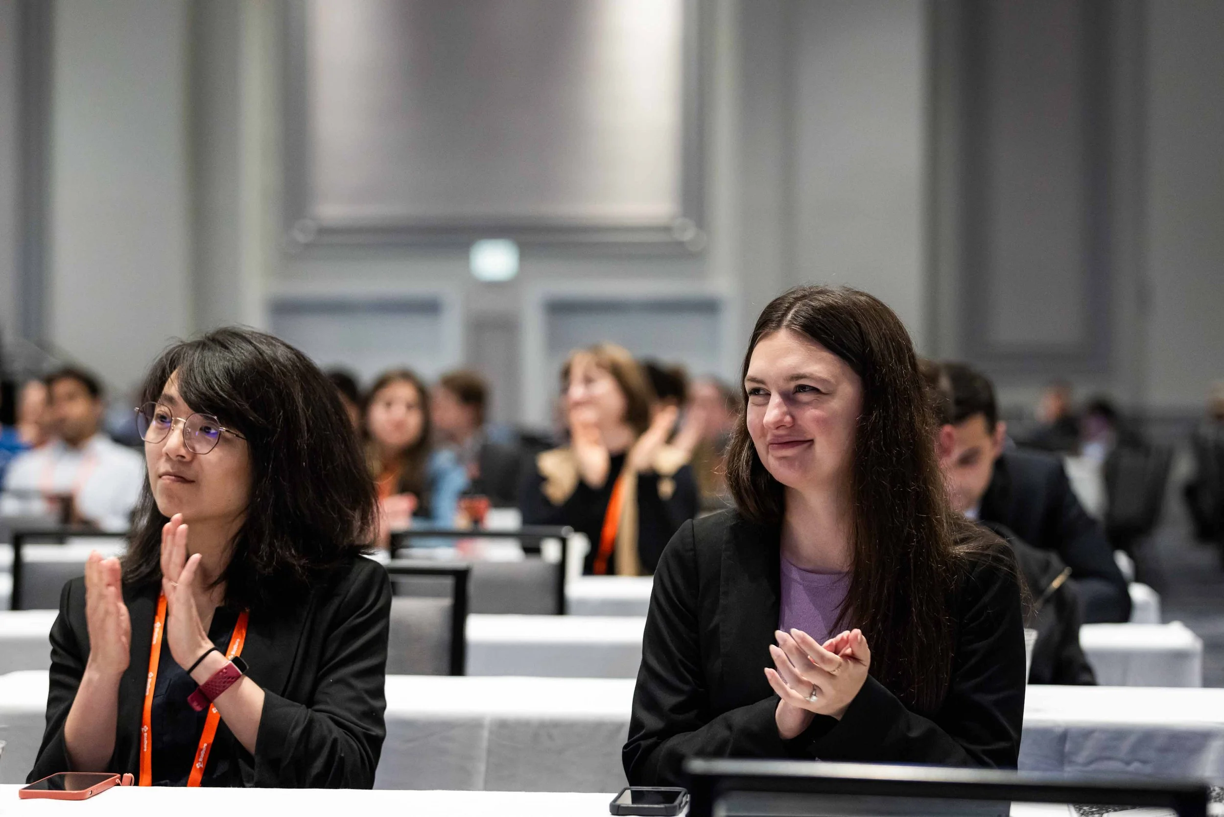 Two women sitting at a conference table, clapping and smiling, with several people in the background in a formal conference room.