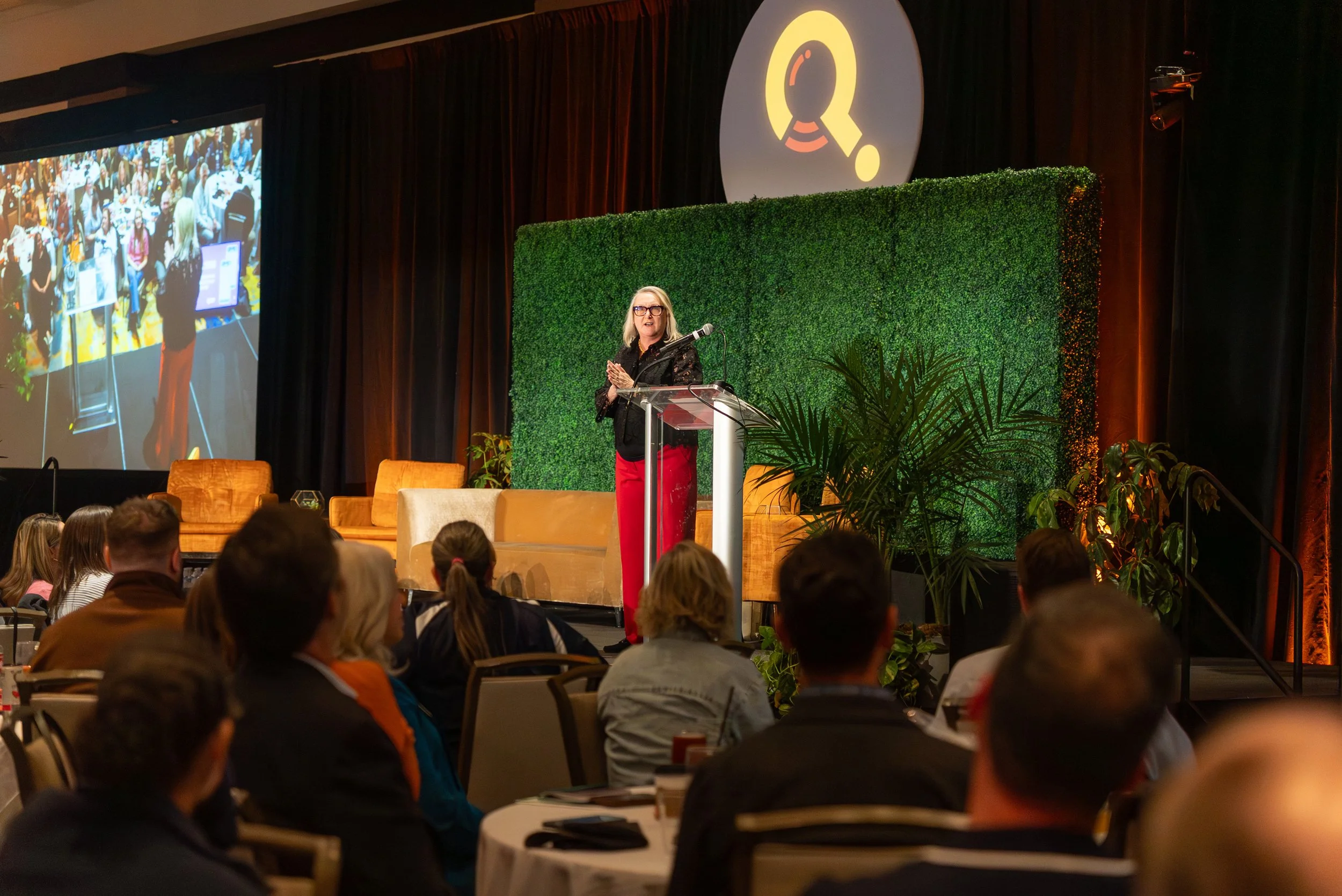 A woman giving a presentation on stage at a conference with an audience seated at round tables. The stage decor includes a green hedge wall and plants. There is a large screen showing the event. The backdrop displays a logo with a magnifying glass.