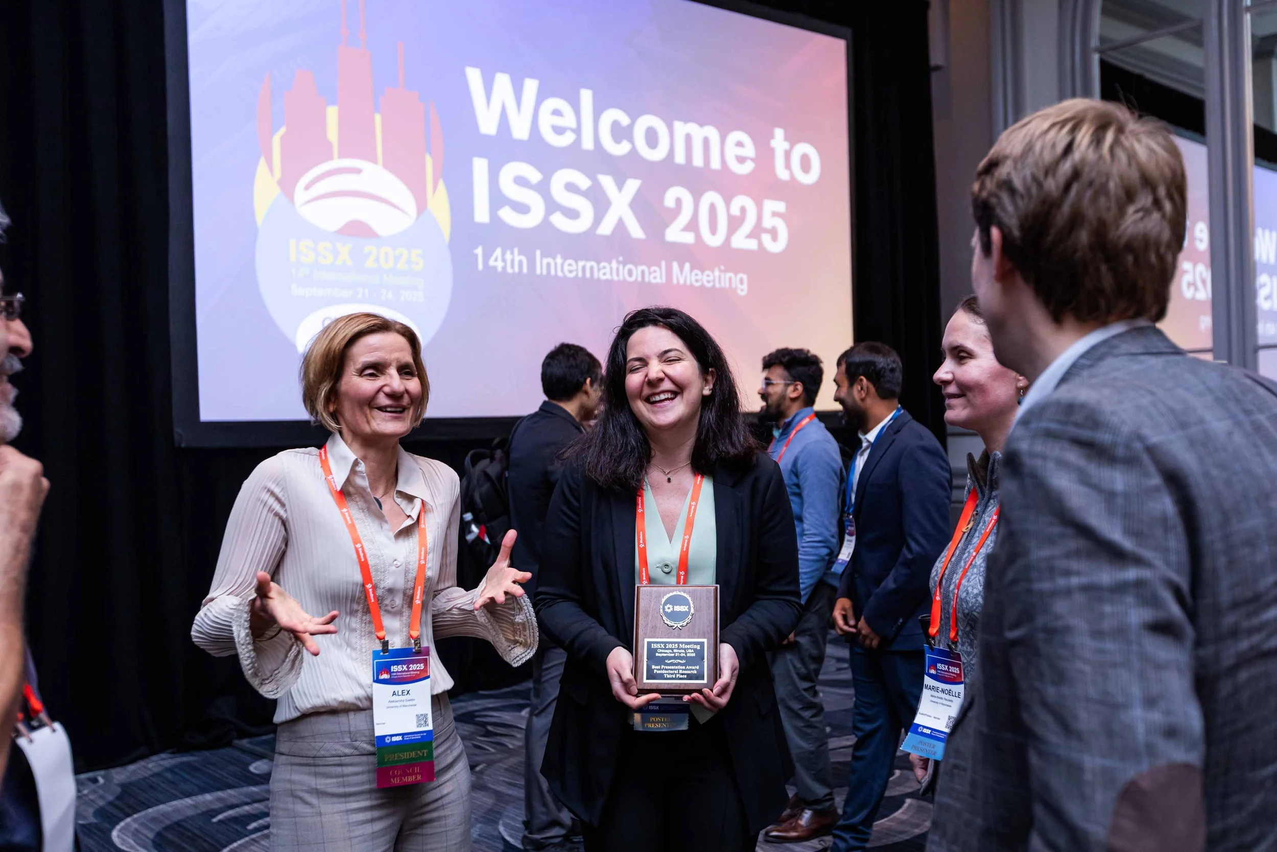Group of people at a conference, smiling and talking in front of a large screen that reads 'Welcome to ISSX 2025, 14th International Meeting.' One woman in the center is holding a plaque, and others are wearing conference badges.