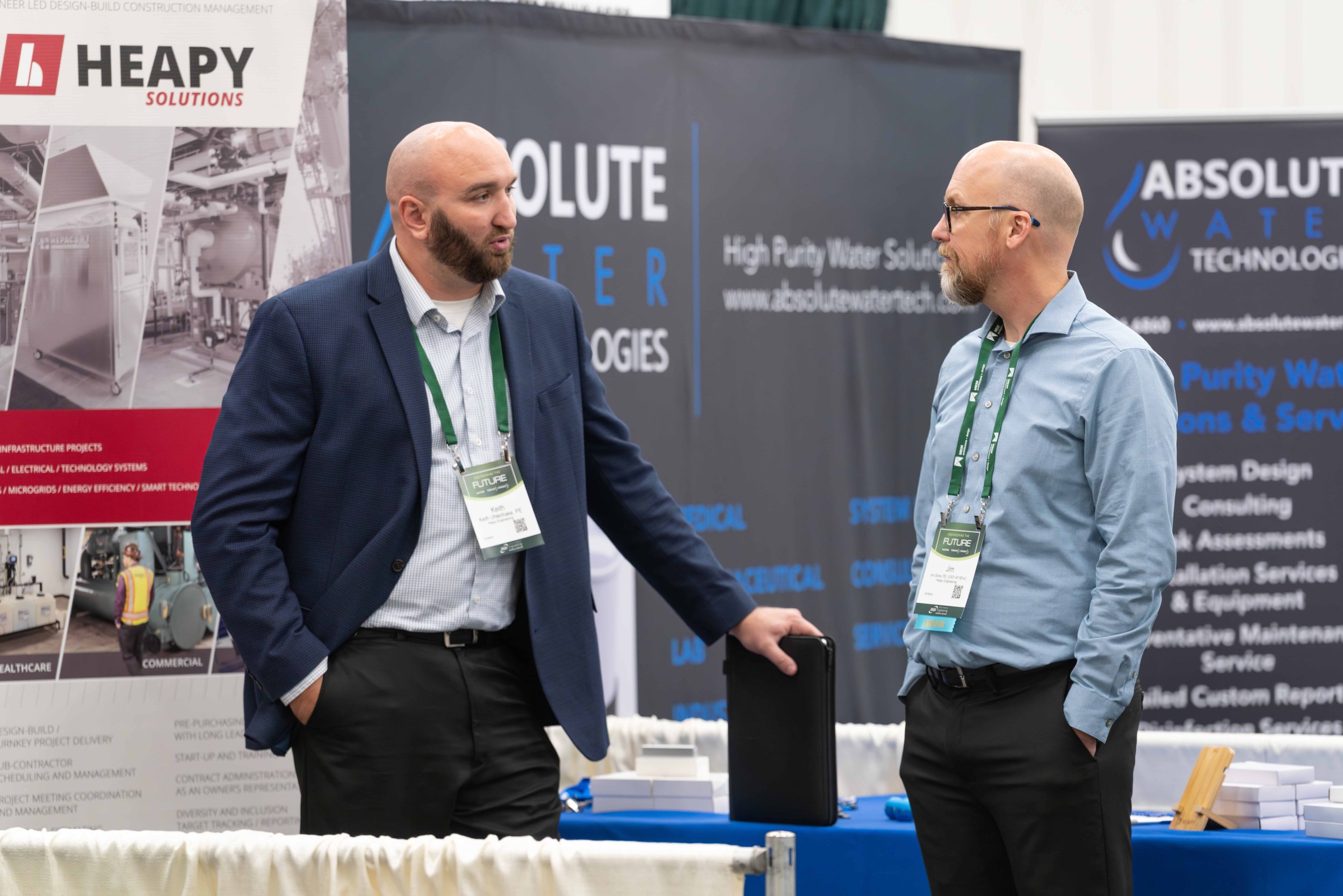 Two men with bald heads and beards engaged in conversation at a trade show booth. One wears a dark blue suit jacket and light blue shirt, and the other wears a light blue shirt. Both have green lanyards with name badges. The background features banne