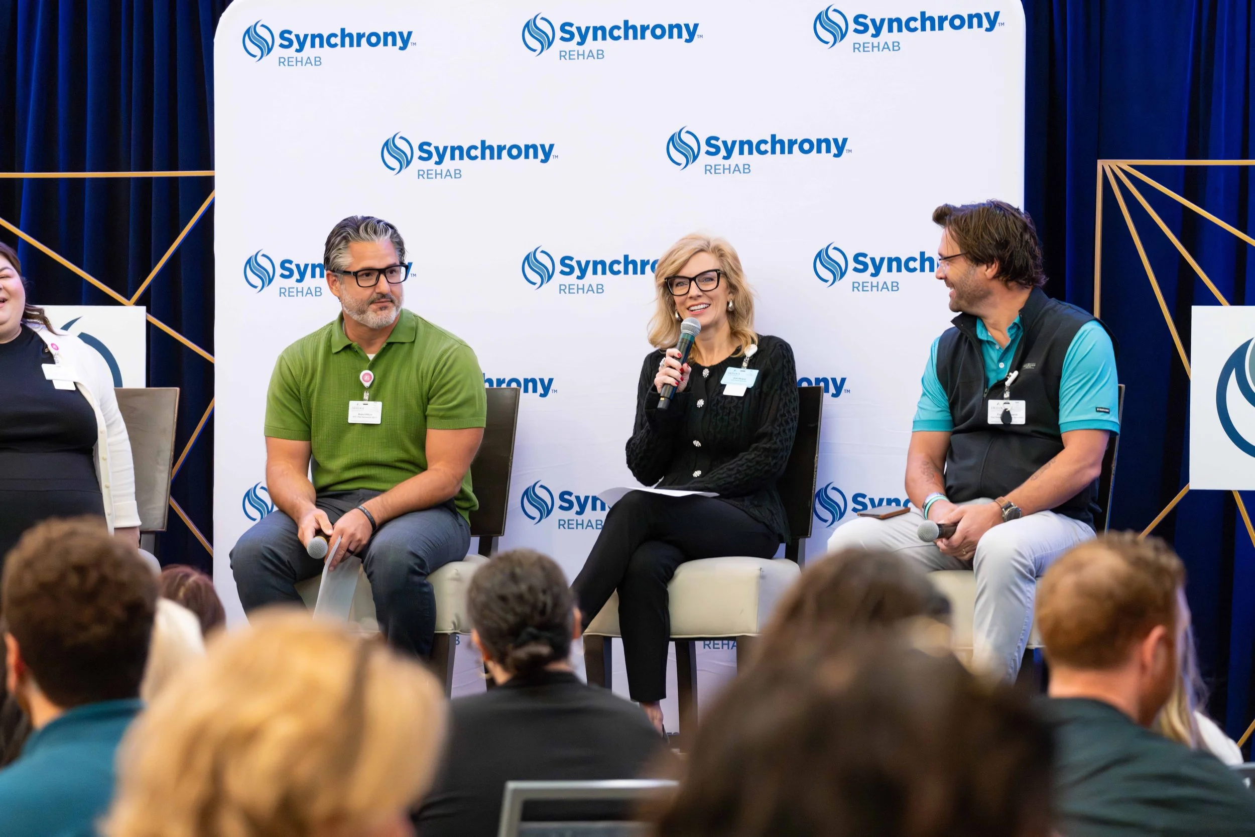 Panel discussion at a social event with three people sitting on stage, speaking into microphones, with a 'Synchrony Rehab' branded backdrop, and audience members seated in the foreground.