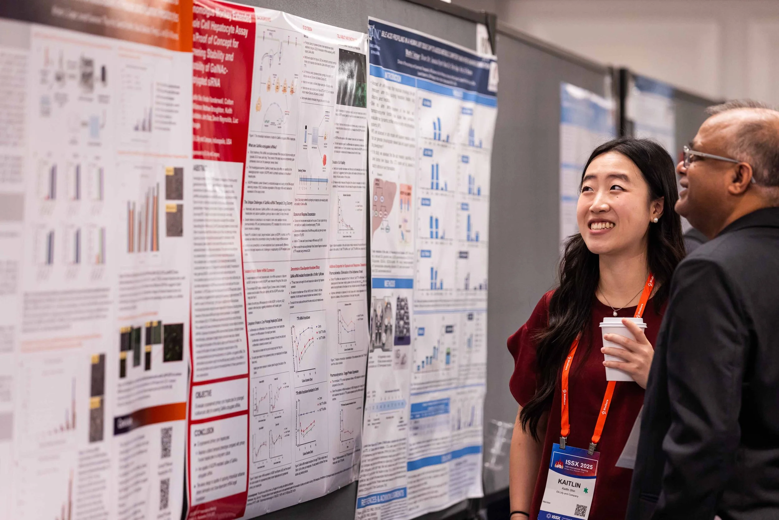 Two people, a woman and a man, are standing in front of scientific research posters at a conference, smiling and engaging in conversation. The woman is holding a coffee cup.