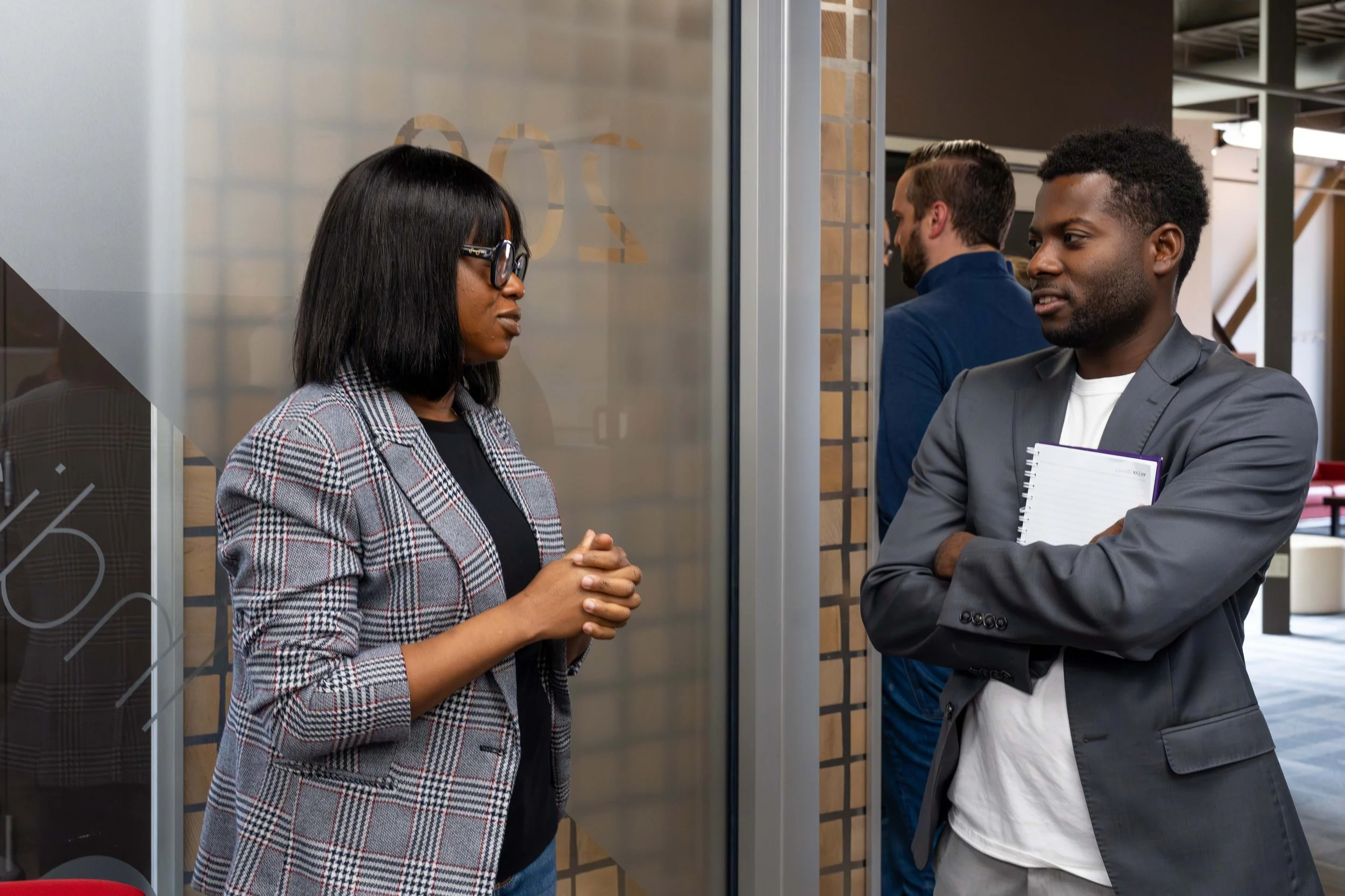 Two business professionals, a woman with black hair and glasses wearing a plaid blazer and a man with short hair and a beard in a grey suit, are having a conversation in an office hallway. They are standing on either side of a glass wall.