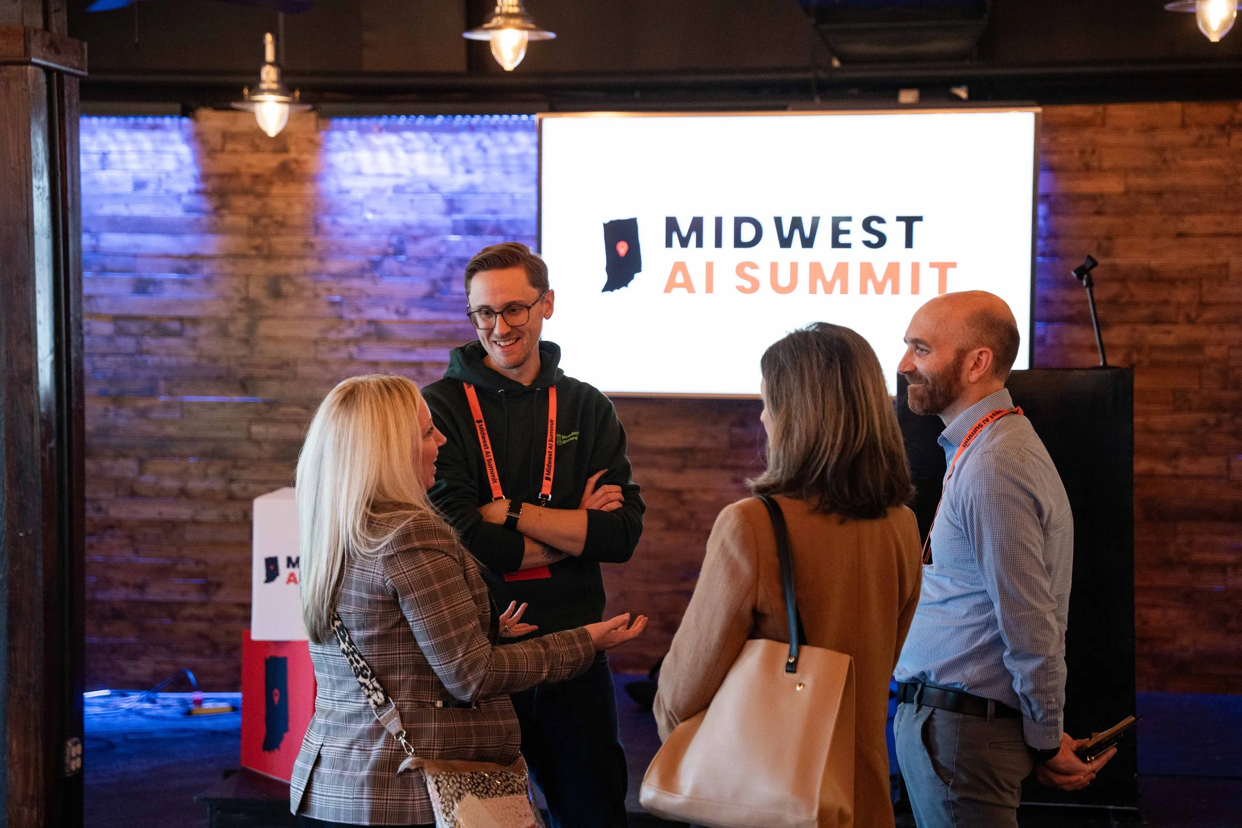 Four people are standing and talking in front of a large screen at the Midwest AI Summit, with a wood-paneled wall background and hanging lights above.