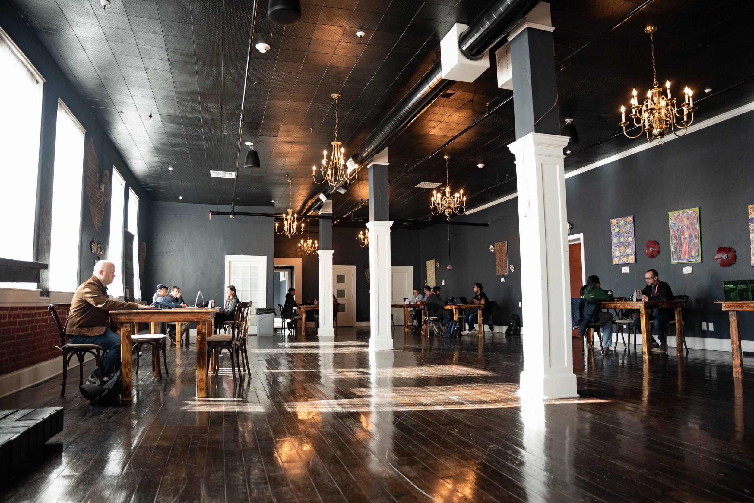 A spacious, well-lit room with dark walls, hardwood floors, and chandeliers hanging from the ceiling. Several tables are occupied by people working or studying, with artwork hanging on the walls and large windows letting in natural light.