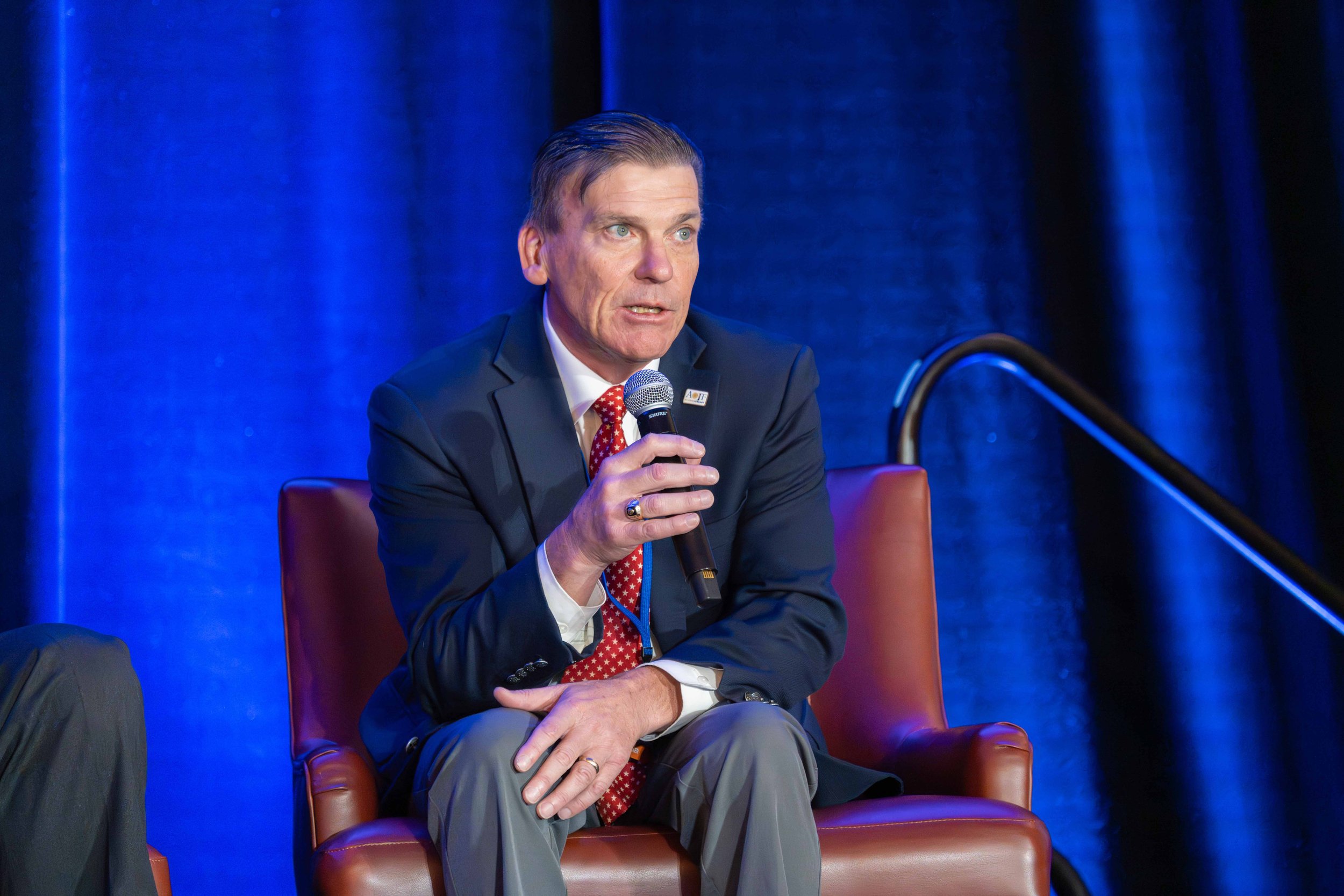 A man in a dark suit, white shirt, and red polka dot tie speaking into a microphone while seated on a stage with blue curtains.