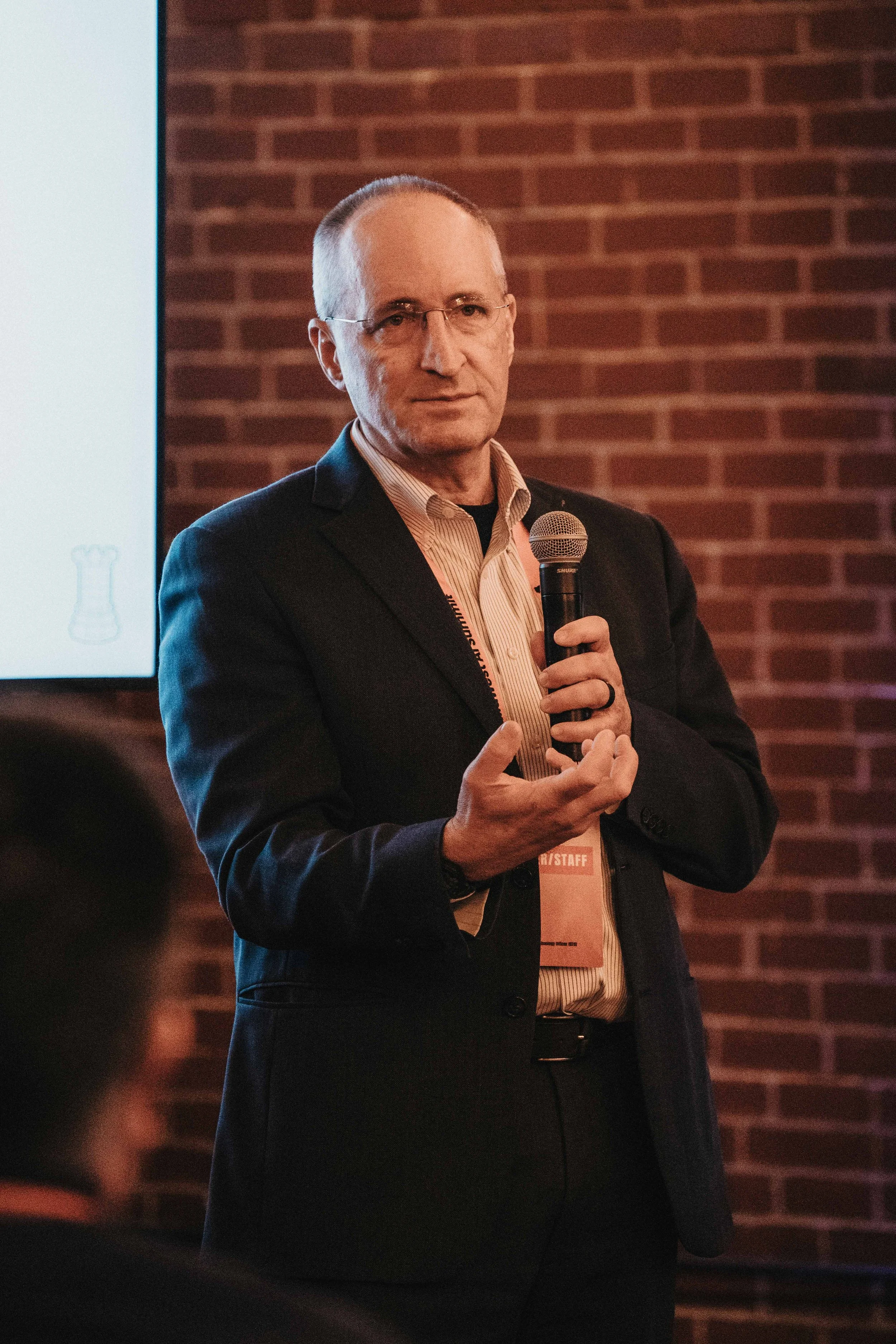 A man in a suit holding a microphone, giving a presentation in front of a brick wall.