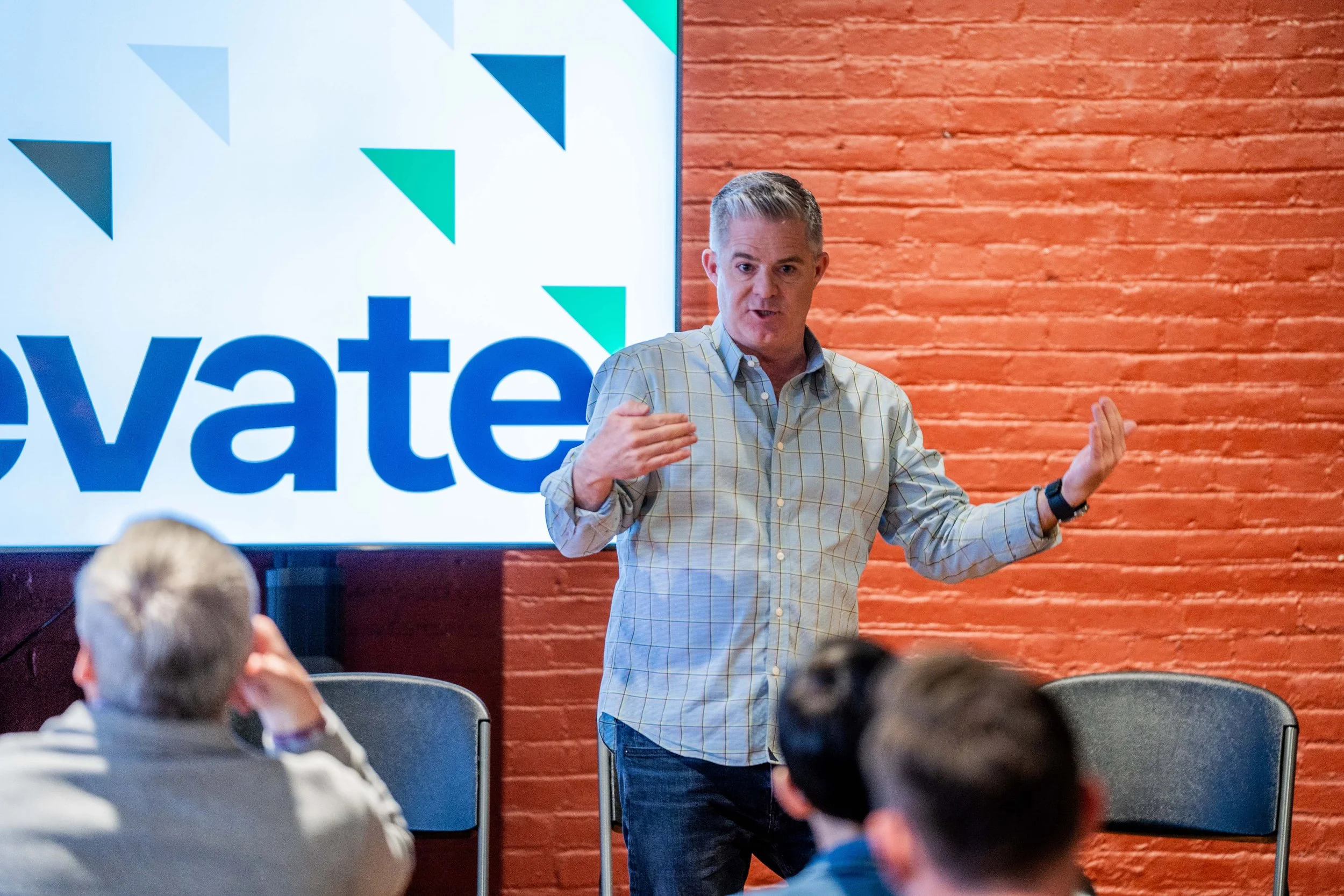 Man giving a presentation in front of a screen displaying the word 'elevate' with a multicolored geometric design, in a room with a red brick wall