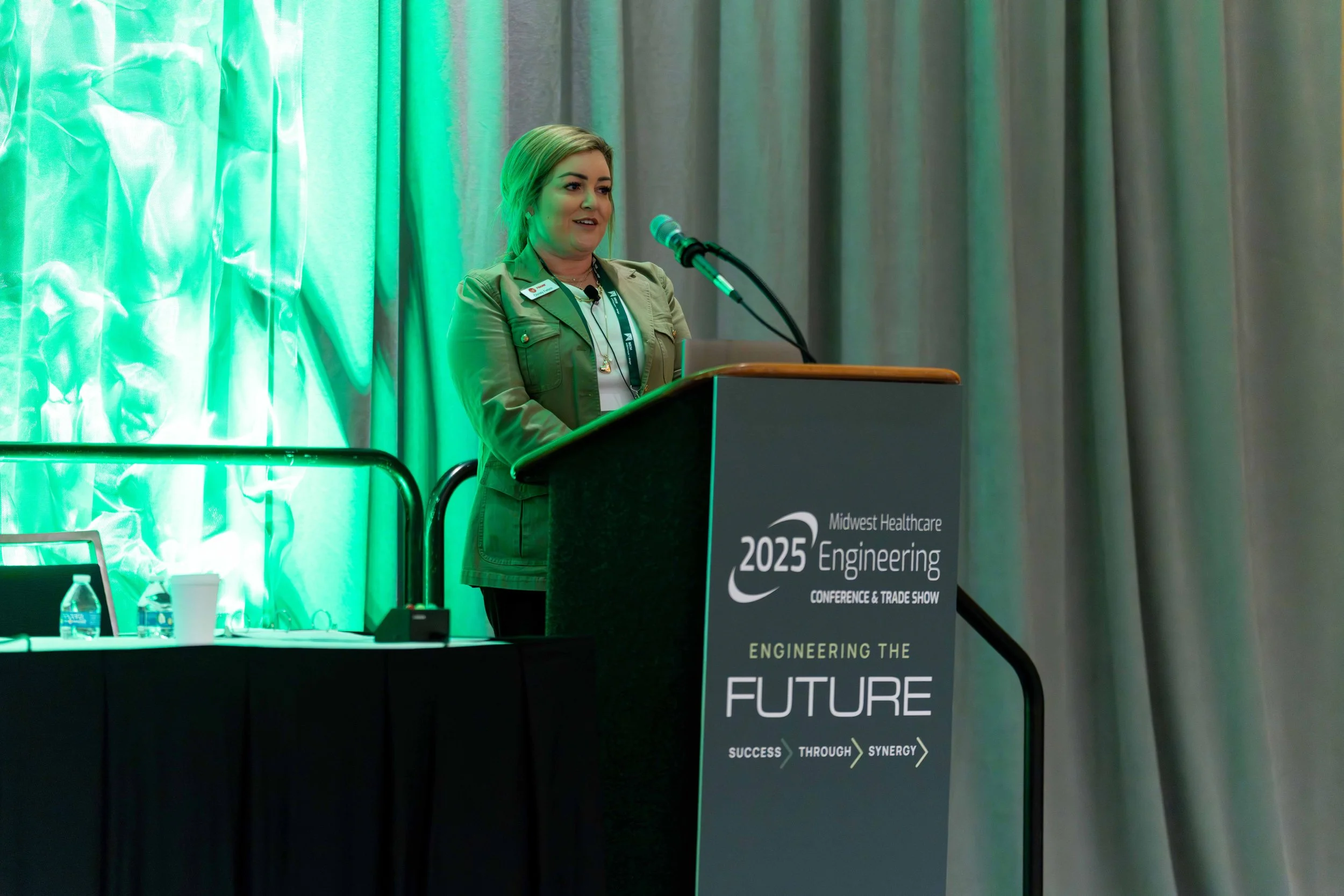 A woman standing at a podium speaking at a conference, with a backdrop featuring green lighting and gray curtains. The podium displays a sign for Midwest Healthcare Engineering 2025 Conference & Trade Show, with the slogan "Engineering the Future" an