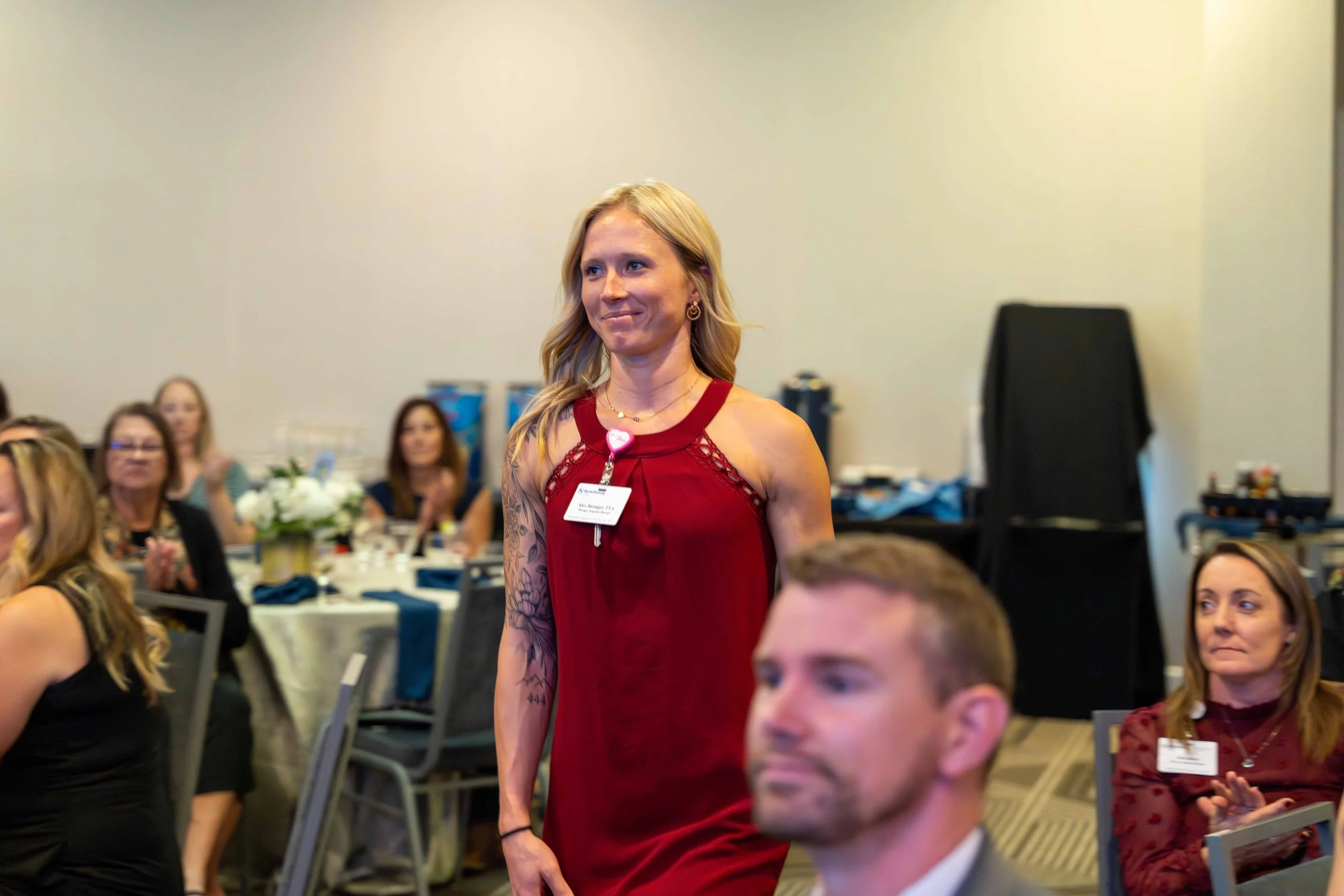 A woman in a red dress standing at a conference or event, with people seated at tables clapping and watching in the background.