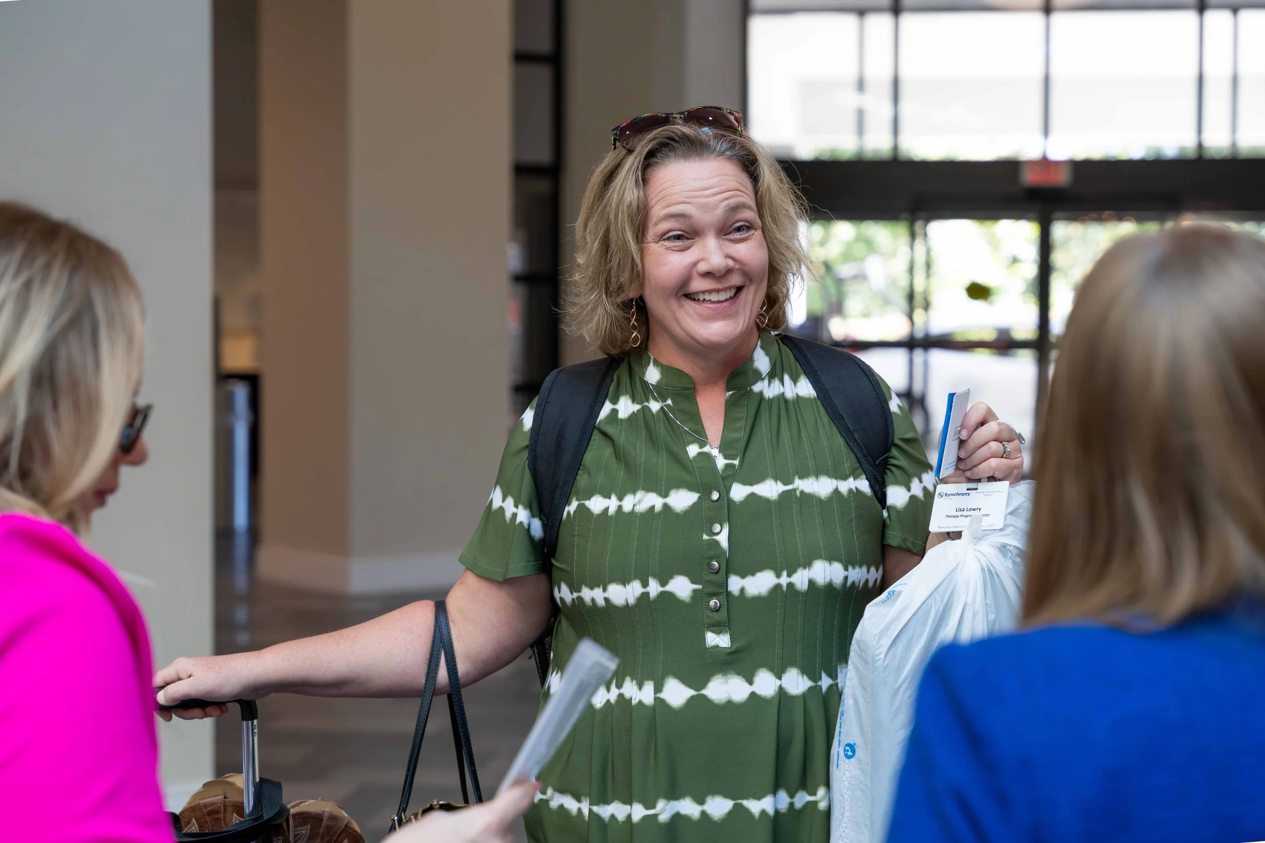 A smiling woman with curly blonde hair, wearing a green and white tie-dye dress, sunglasses on her head, and a black backpack, speaks with two women at what seems to be a hotel lobby or reception area.
