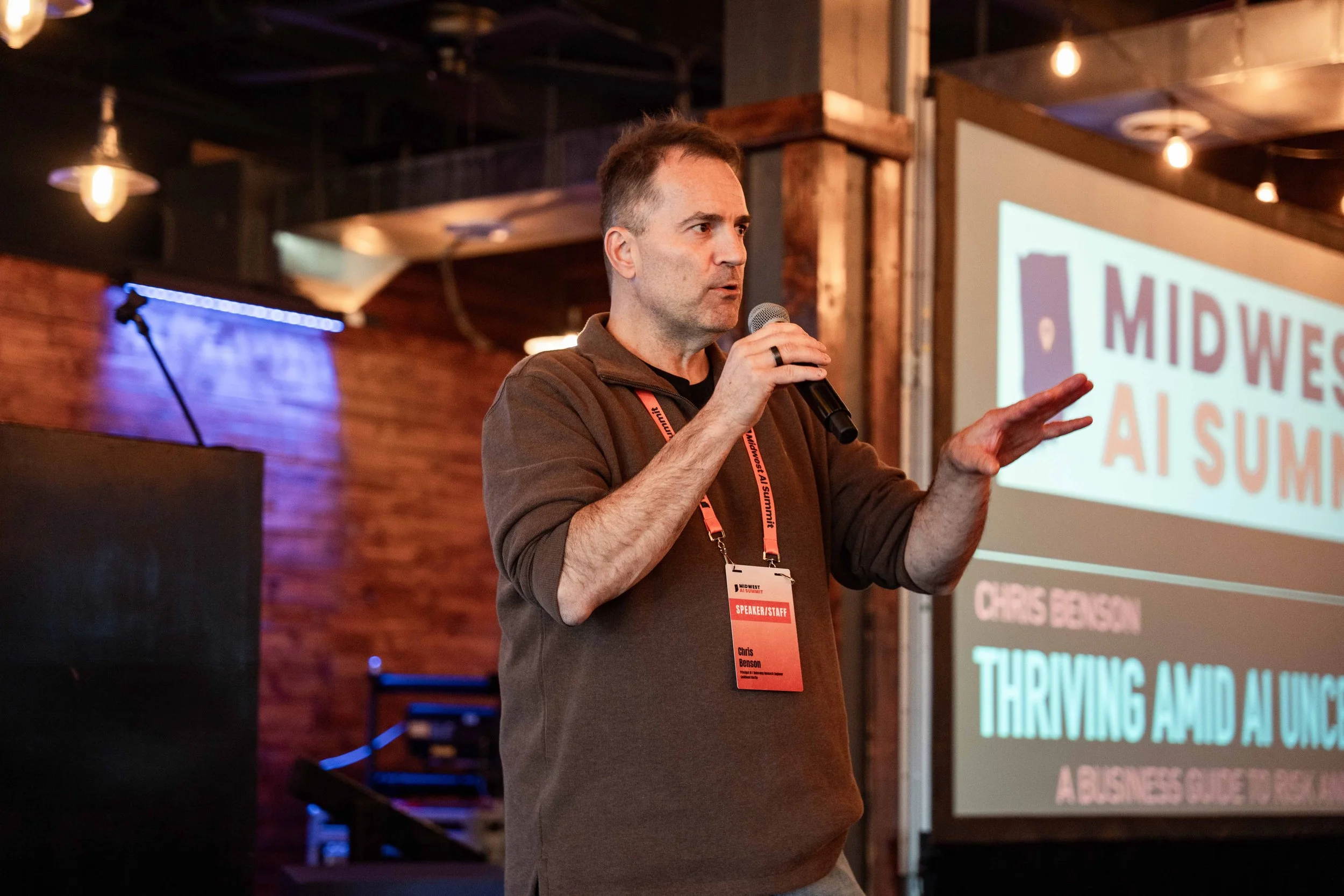 A man in a brown sweater speaking into a microphone at a conference, with a projector screen behind him displaying the title 'Midwest AI Summit: Thriving Amid Uncertainty'.