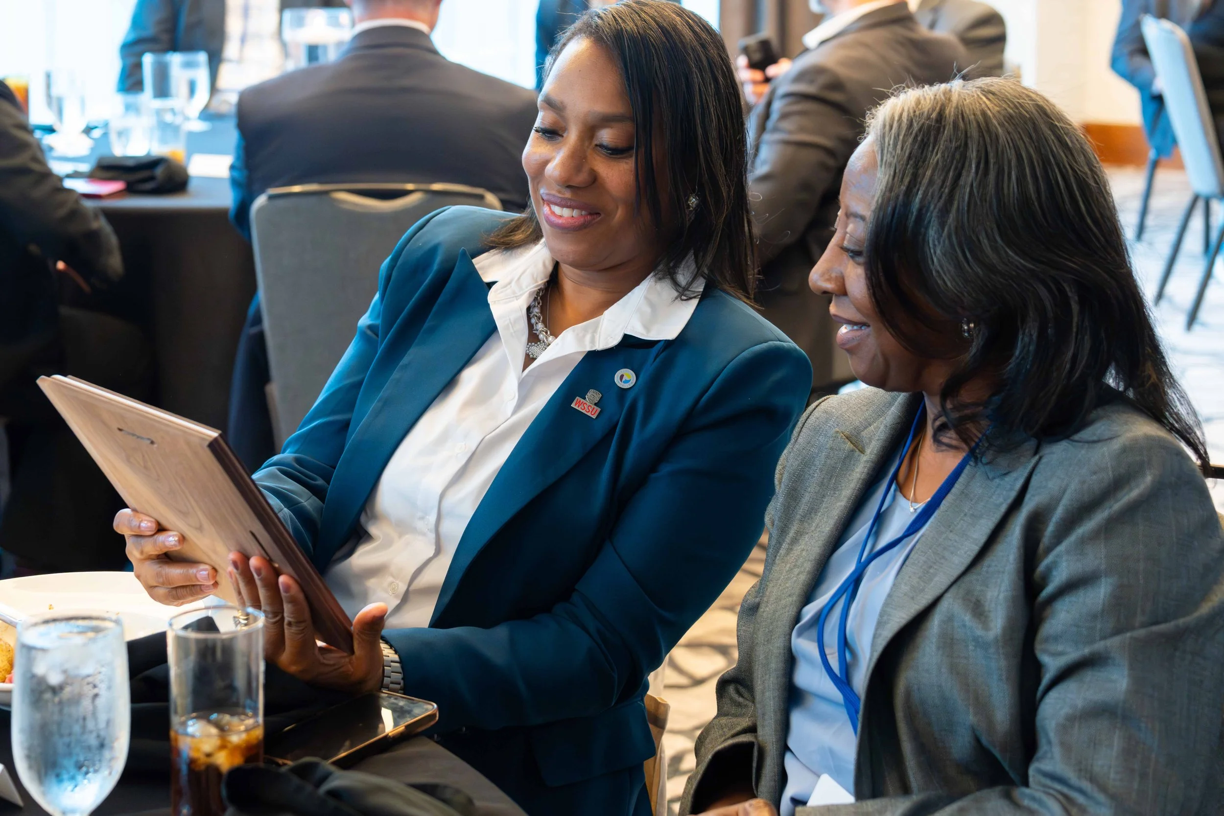 Two women sitting at a table, looking at a tablet. They are dressed in business attire, smiling, and appear to be at a professional conference or meeting.