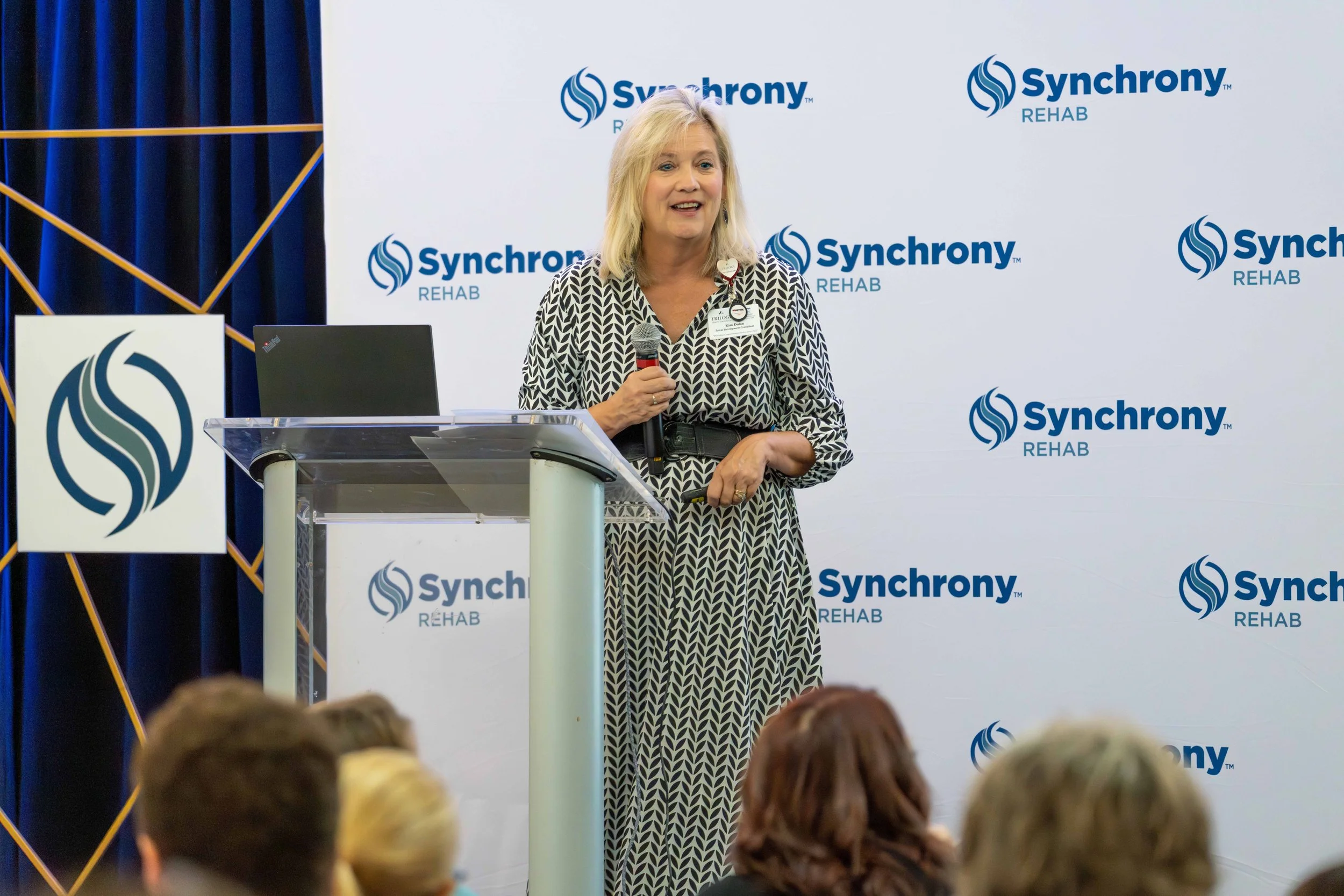 Woman giving a presentation at an event for Synchrony Rehab, standing behind a clear podium with a laptop on it, holding a microphone, with an audience watching.