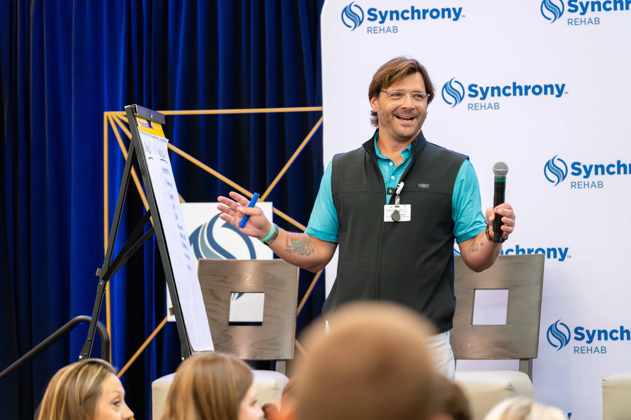 Man giving a presentation at Synchrony Rehab event with a whiteboard, holding a microphone in one hand and a marker in the other, standing in front of a backdrop with Synchrony Rehab logos, with audience members visible in the foreground.