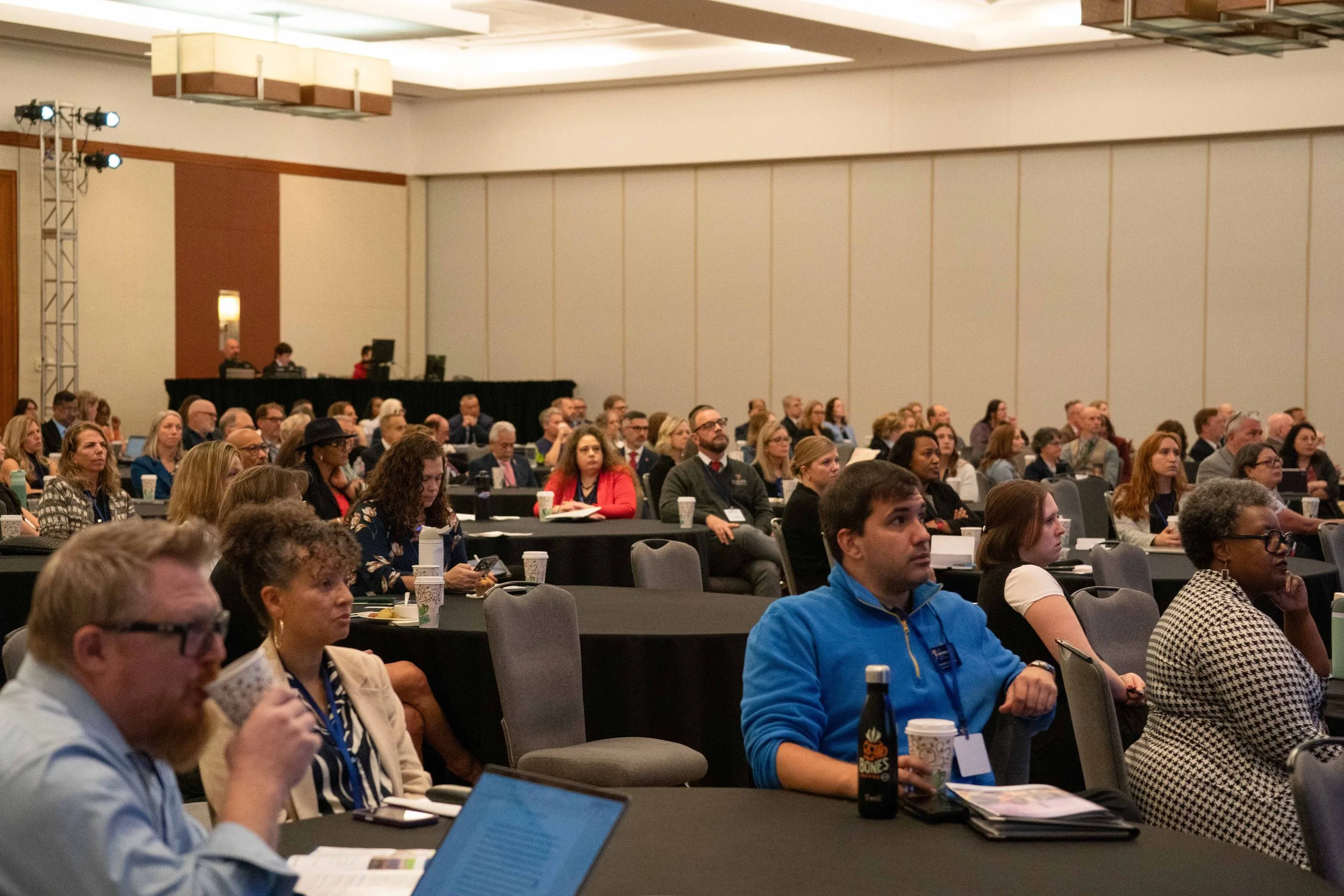 Audience attending a conference or seminar in a large, well-lit room, sitting at round tables with laptops, notebooks, and beverages.