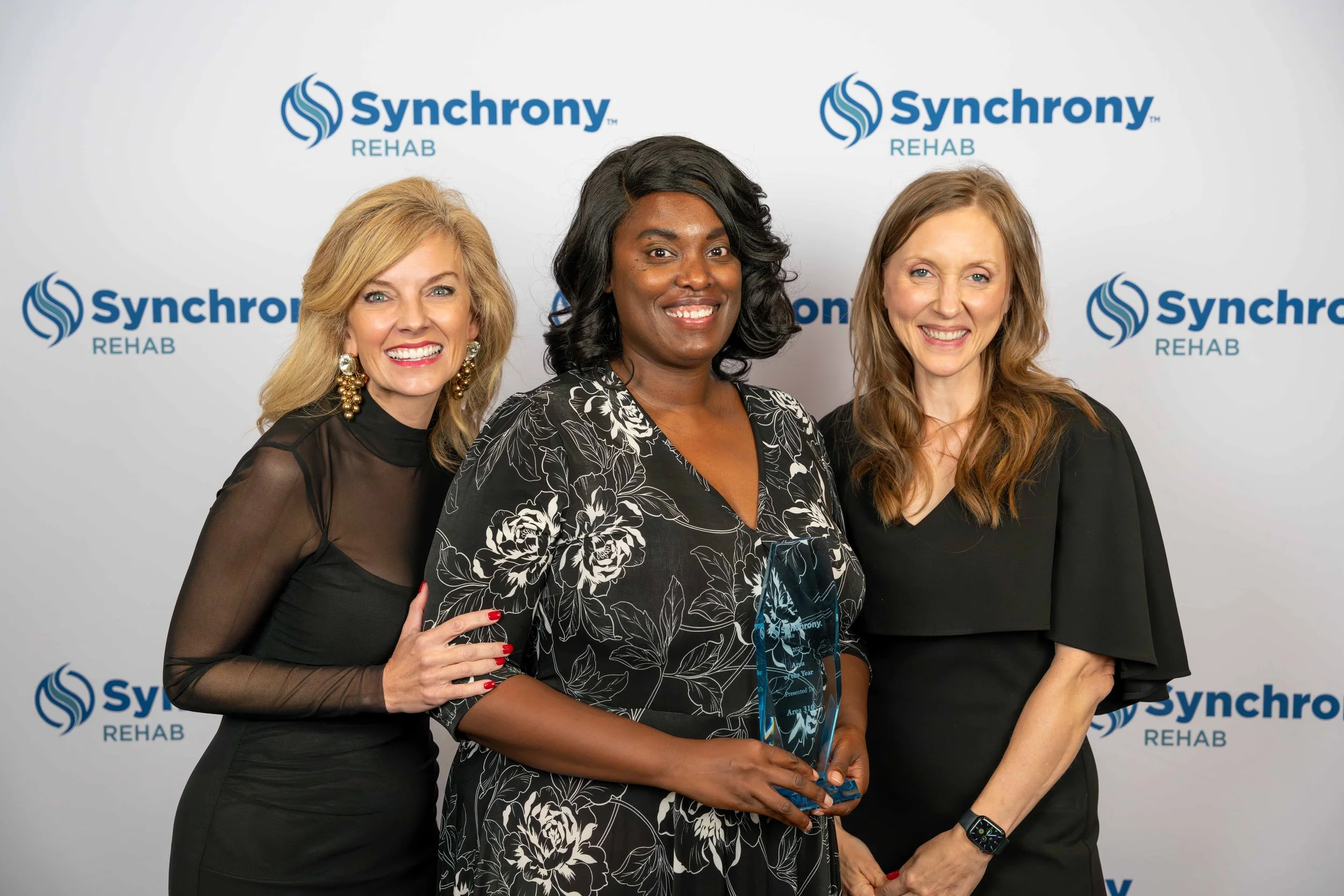 Three women standing together at a reception, one holding an award, in front of a backdrop with "Synchrony Rehab" logos.