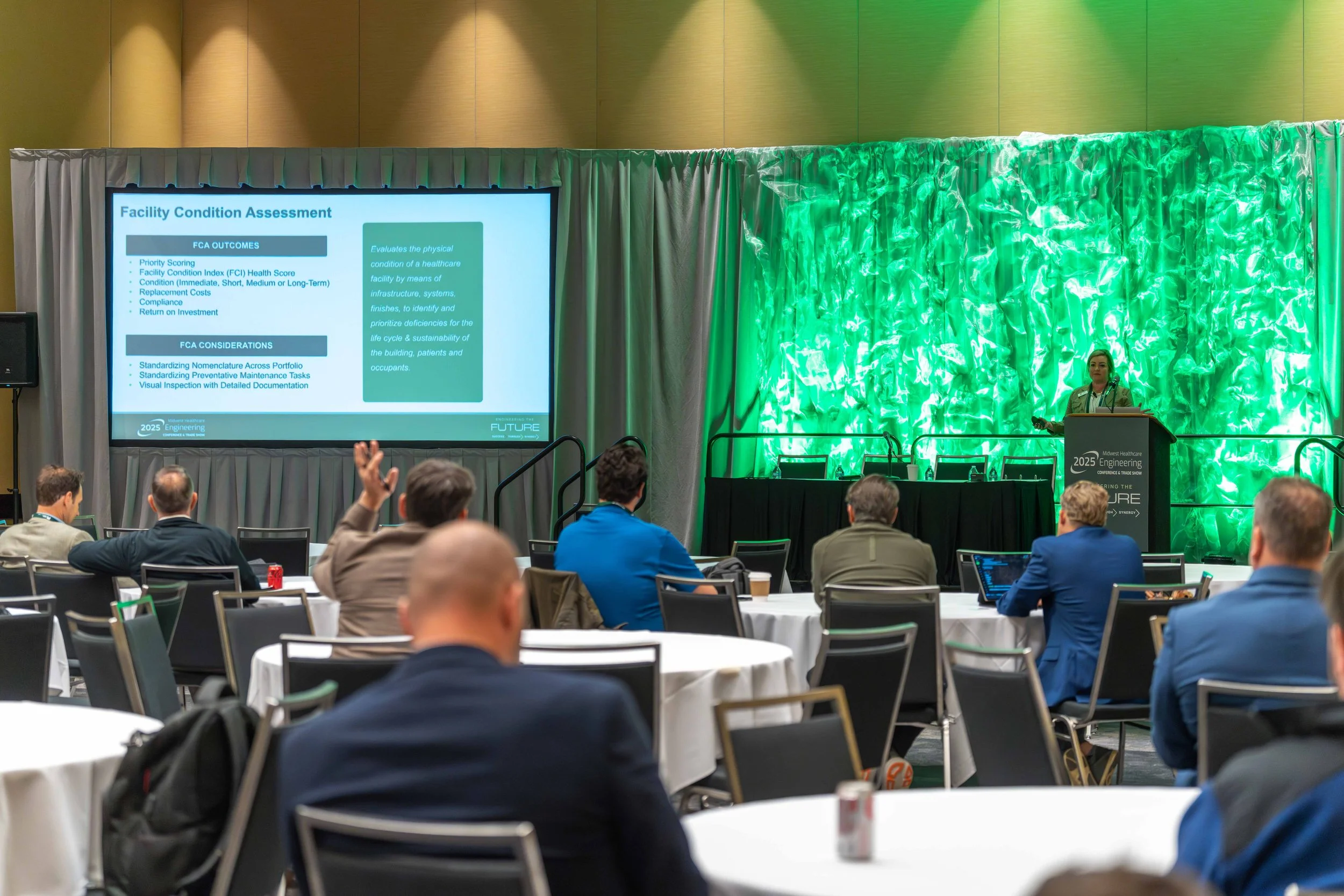 A woman presenting at a conference stage with a large screen displaying a slide titled 'Facility Condition Assessment'. The conference is in a large room with round tables, each with people seated, some with laptops and drinks. The stage backdrop is 