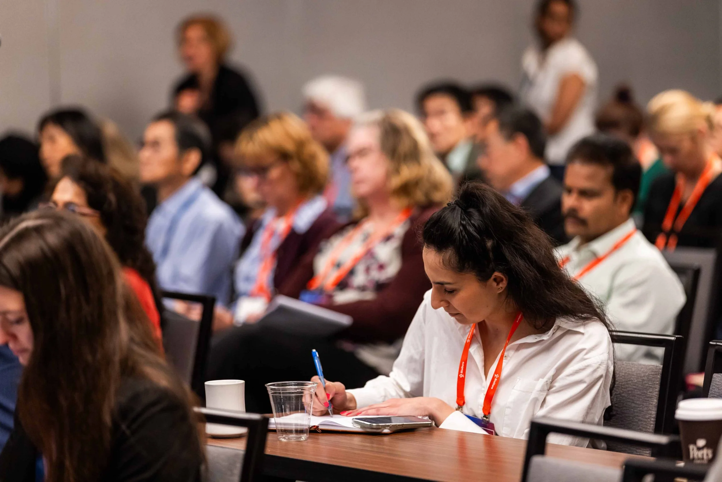 A woman in a white shirt taking notes during a conference, surrounded by an audience of men and women seated at tables in a conference room.