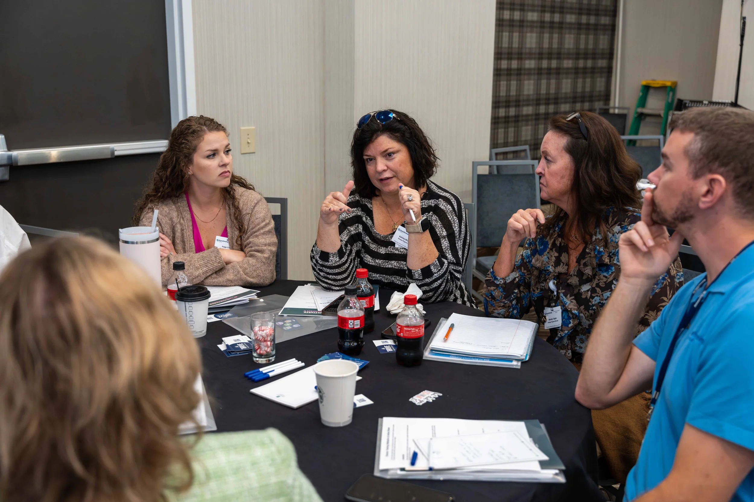 Group of adults at a meeting or workshop engaged in discussion, seated around a table with notebooks, pens, and beverages.