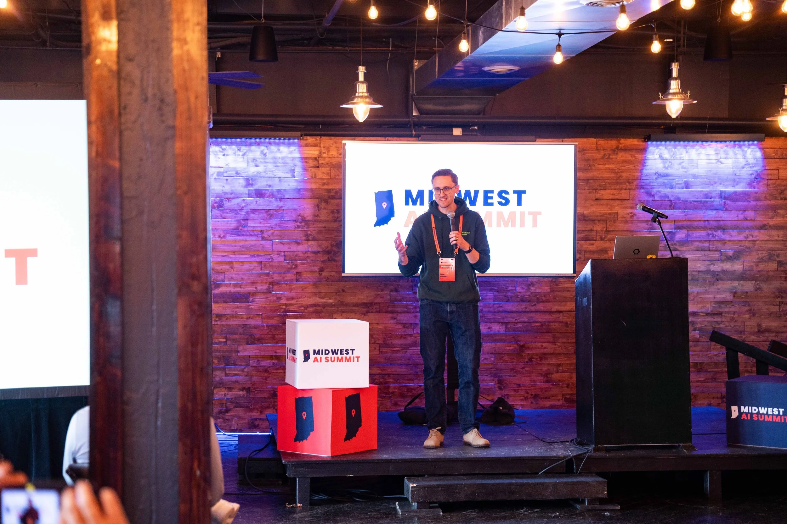 A man giving a presentation on a stage at the Midwest AI Summit. He is holding a microphone and wearing a dark hoodie, jeans, and sneakers. Behind him is a large screen displaying the summit's logo, which includes an outline of the state of Indiana w