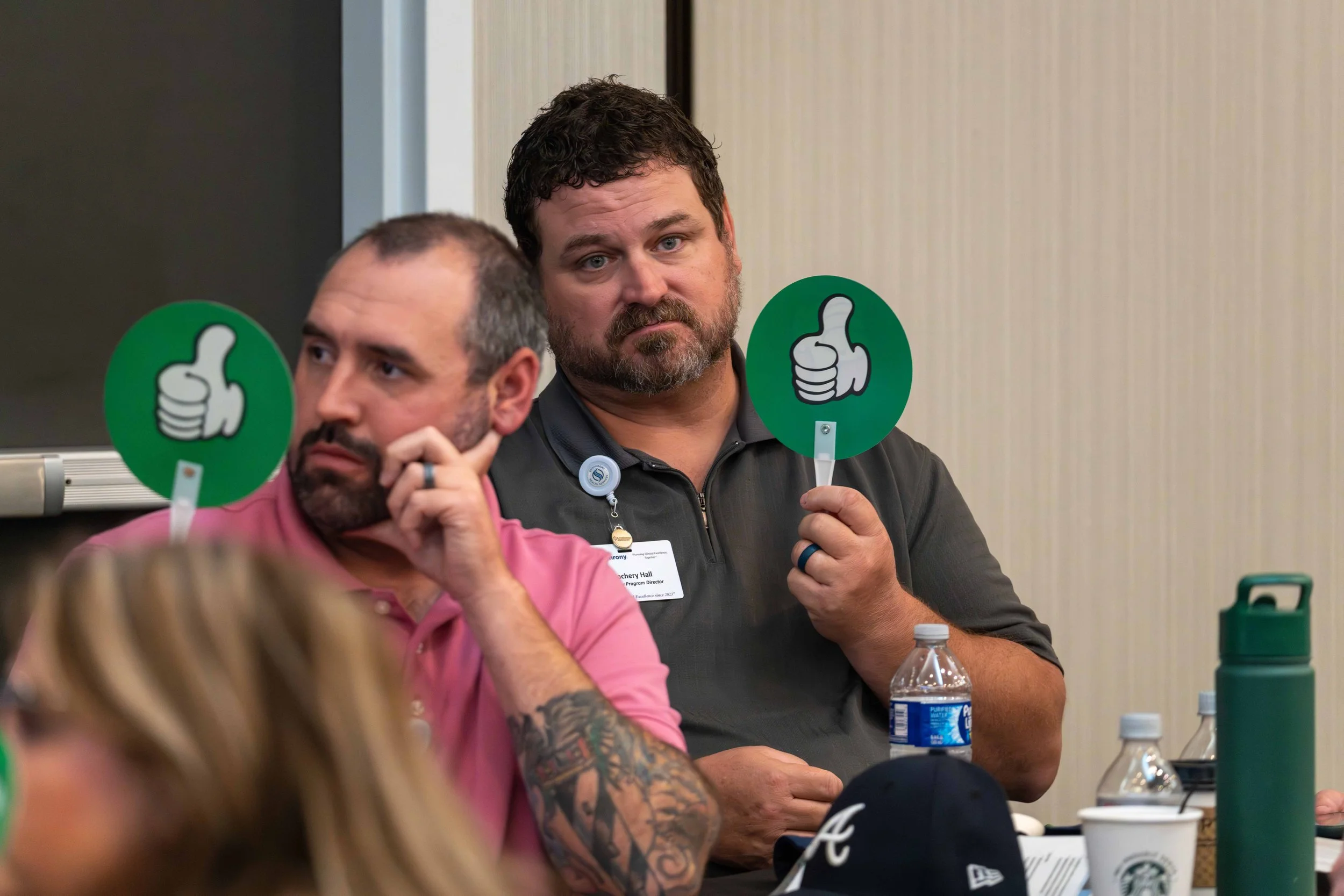 Two men sitting at a table during an event, each holding a green sign with a white thumbs-up symbol. The man in the foreground is wearing a pink shirt and has tattoos on his arm, while the man behind him is wearing a dark gray shirt.