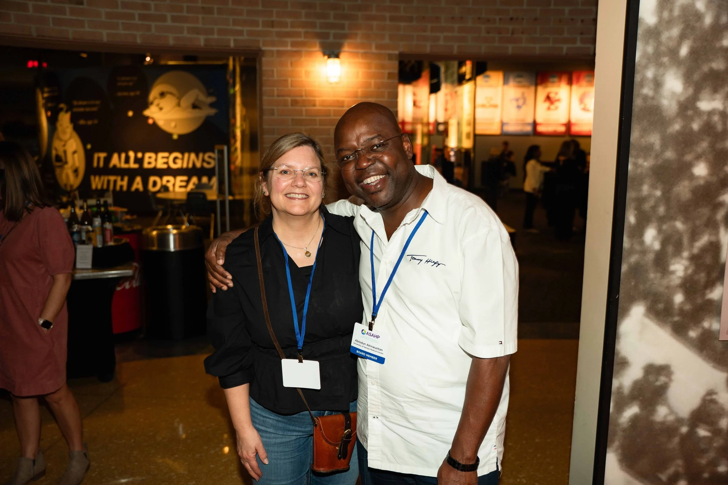 Two smiling people, a woman with glasses and a man, are standing together with their arms around each other at an indoor event or conference, with a bar and a large sign in the background.