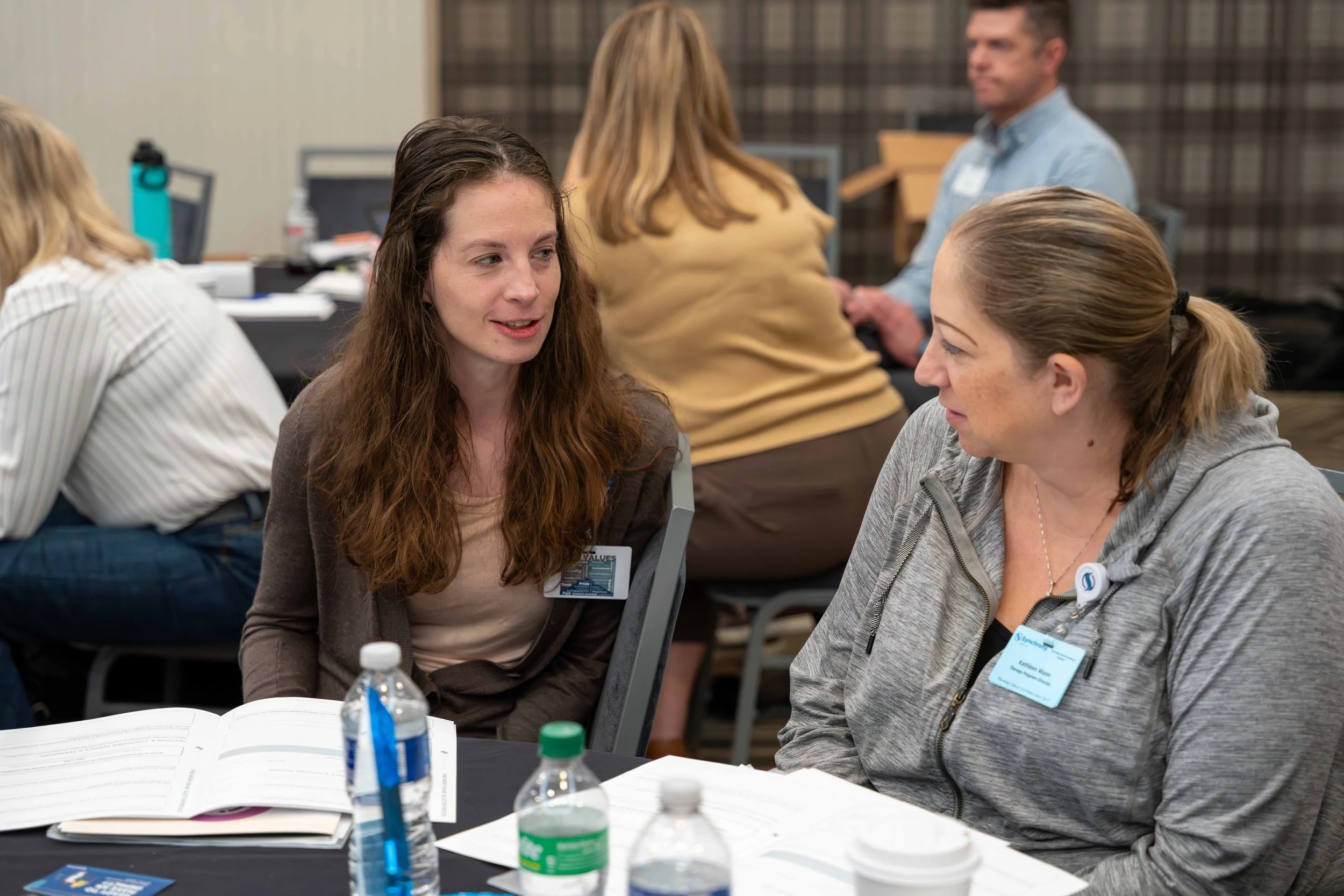 Two women are sitting at a table, engaging in a conversation during a conference or workshop. The table has notebooks, water bottles, and a cup.