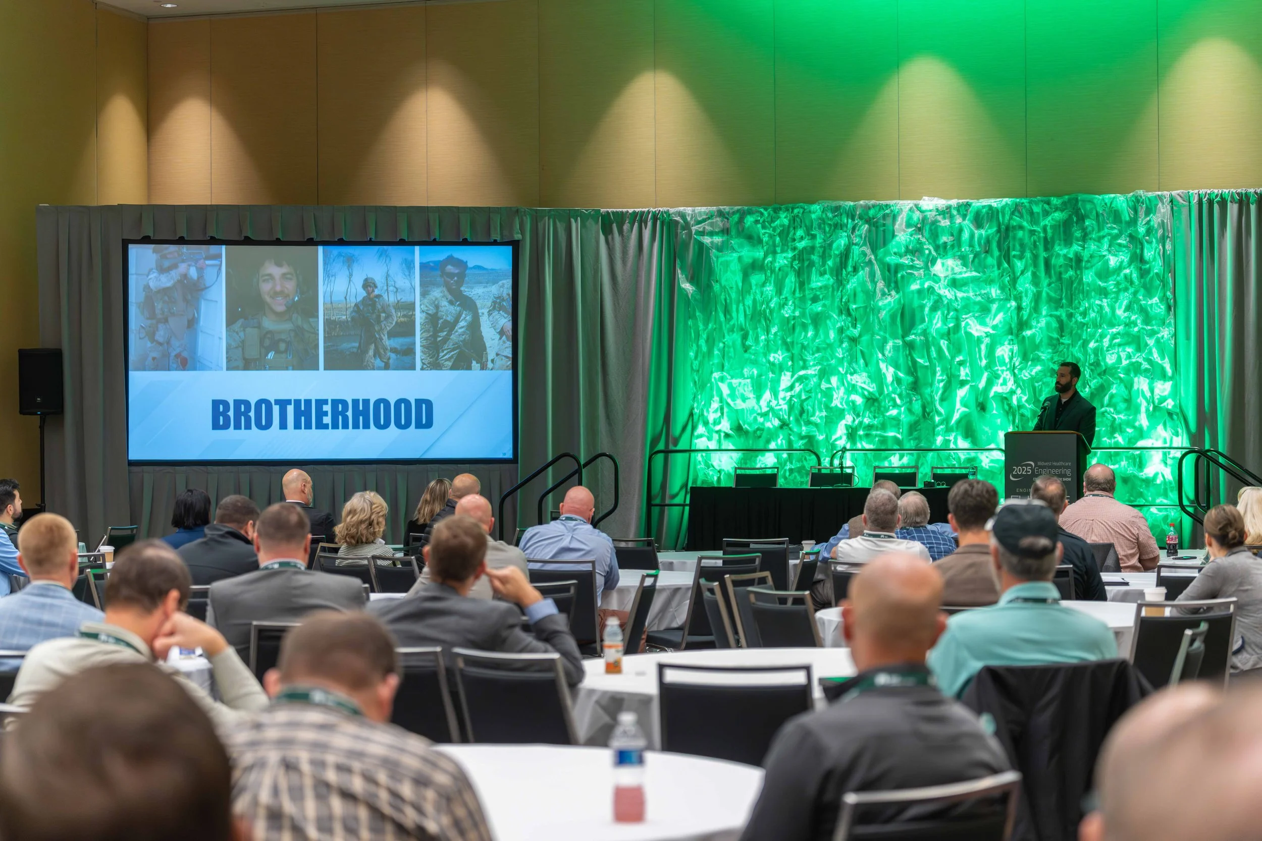 A conference hall filled with people attending a presentation. On the stage, a man stands at a podium, and a large screen displays four photos of a man in military uniform along with the word 'BROTHERHOOD' in bold blue letters. The hall has beige wal