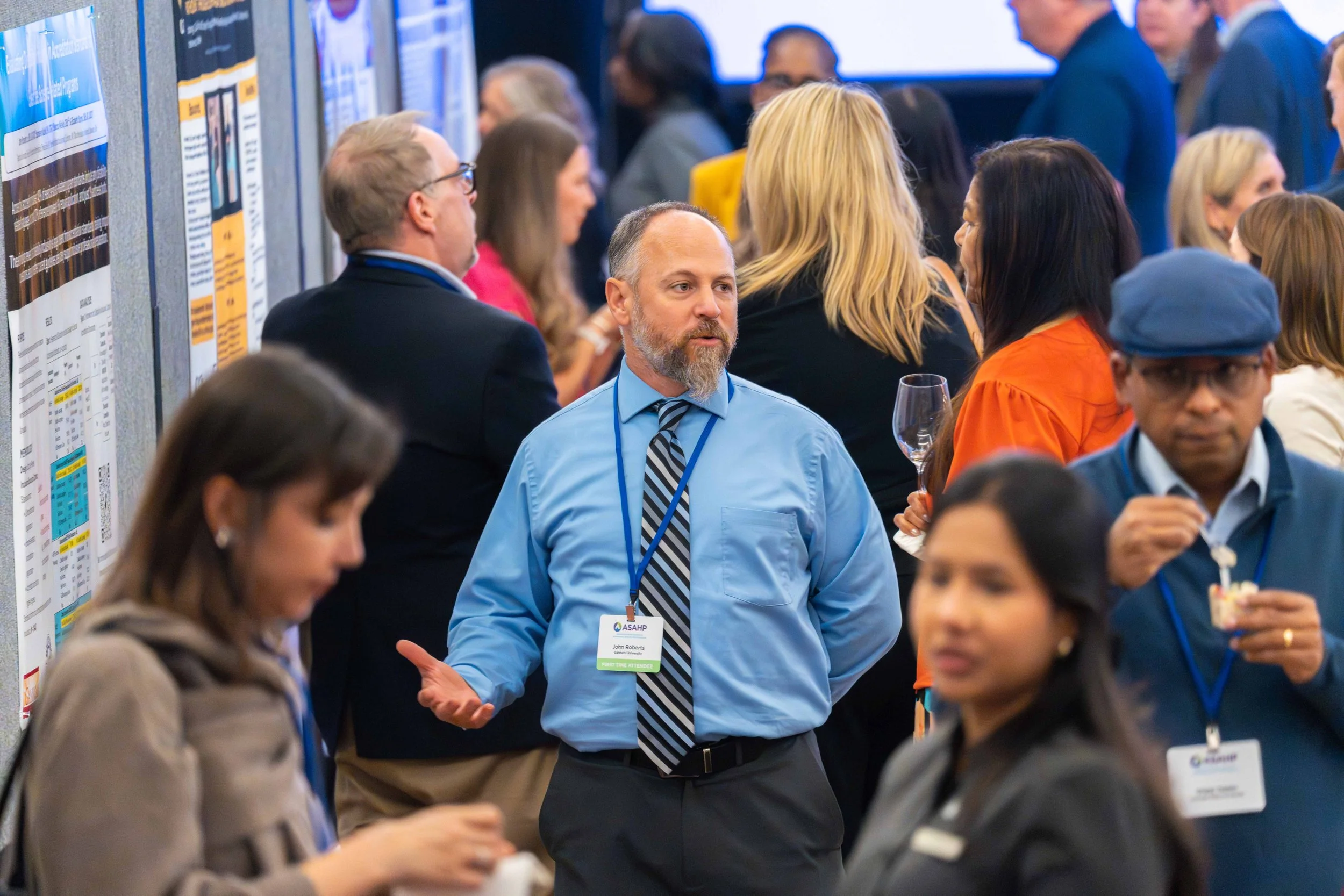 A group of diverse people attending a conference or seminar, standing near informational posters with some engaging in conversation and others consuming refreshments.