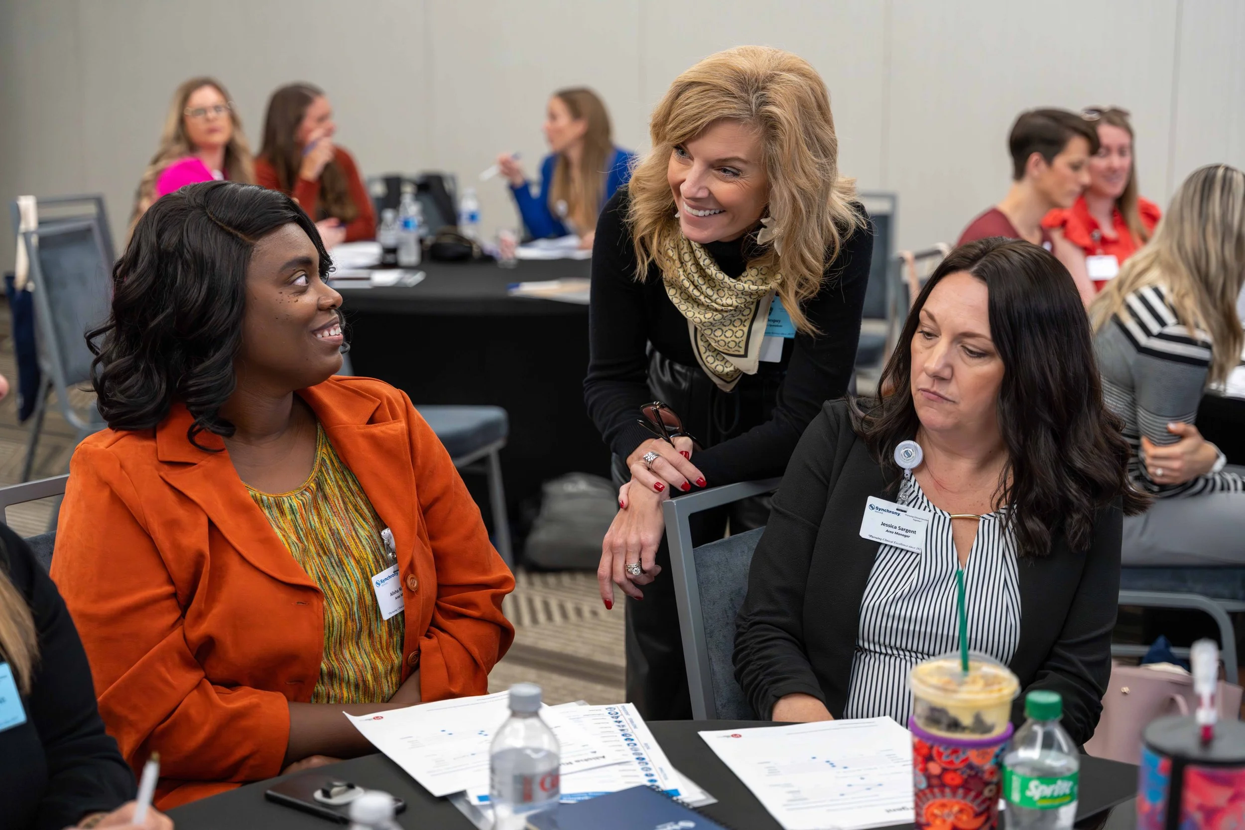Women in a conference room engaging in conversation, with name tags and paperwork on the table.