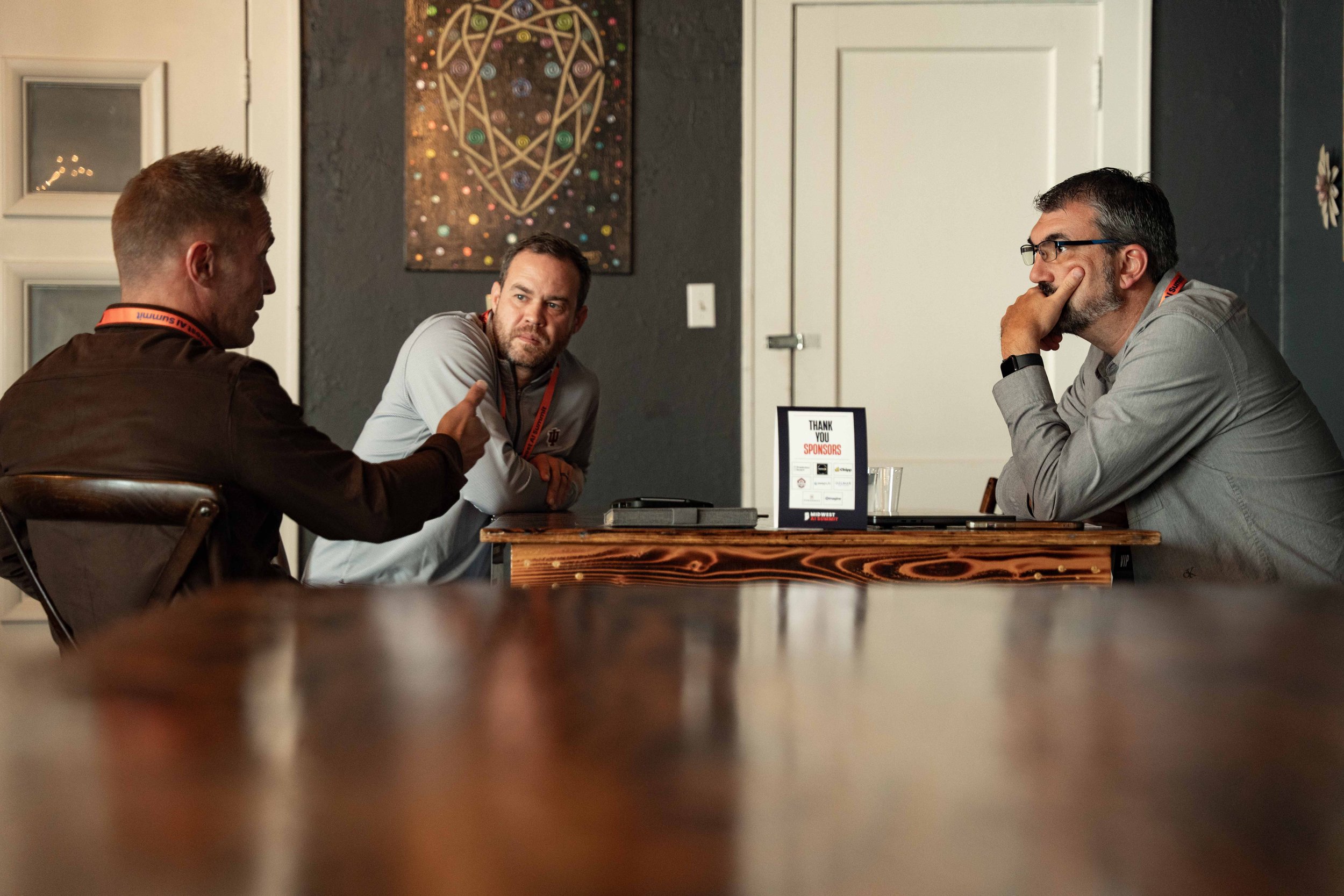 Three men sitting around a table in a meeting or discussion, with one man speaking, the other listening, and one resting his chin on his hand, in a room with dark walls and artwork on the wall.