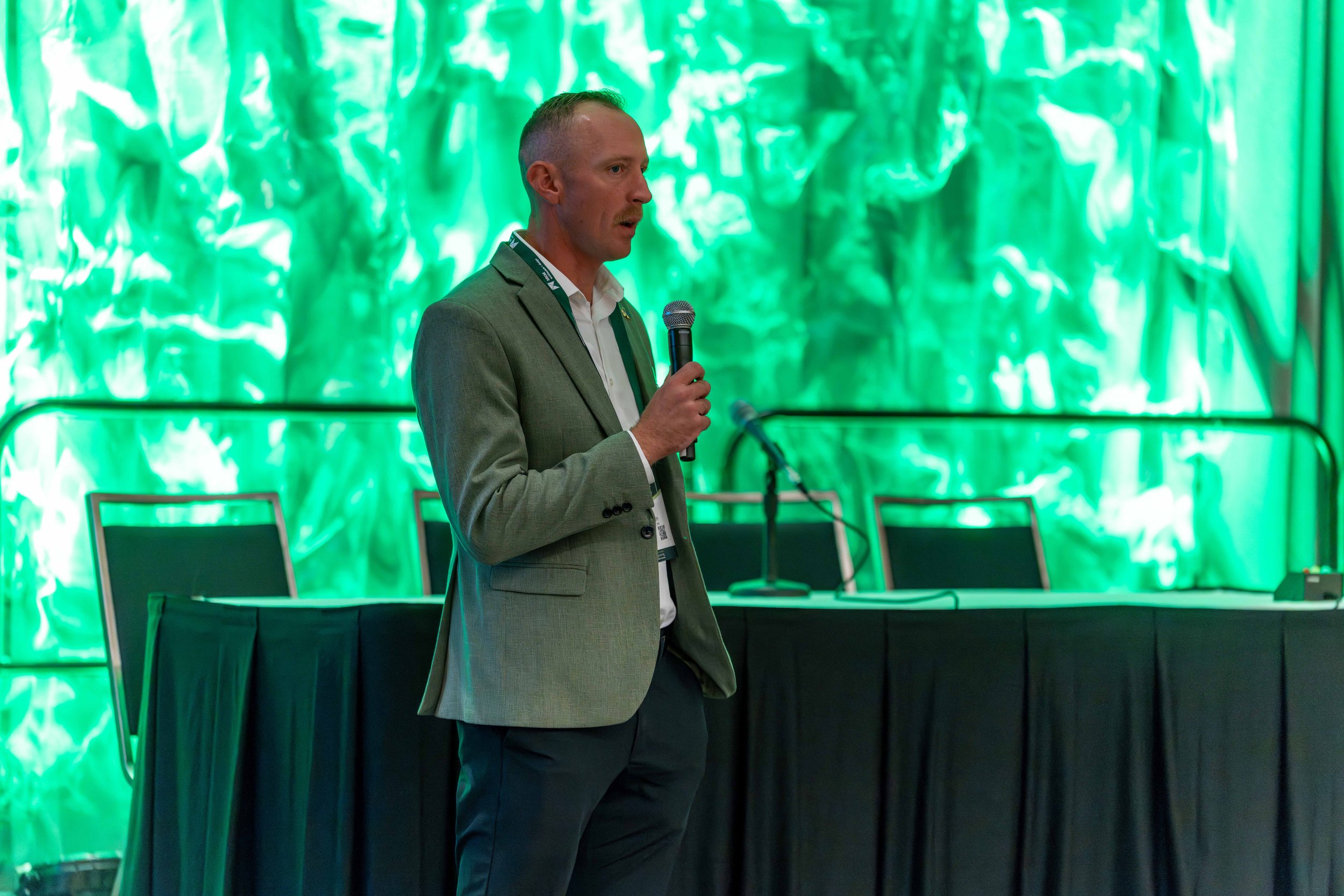 A man in a gray suit and white shirt holding a microphone, standing in front of a green-lit abstract background at a conference or meeting.