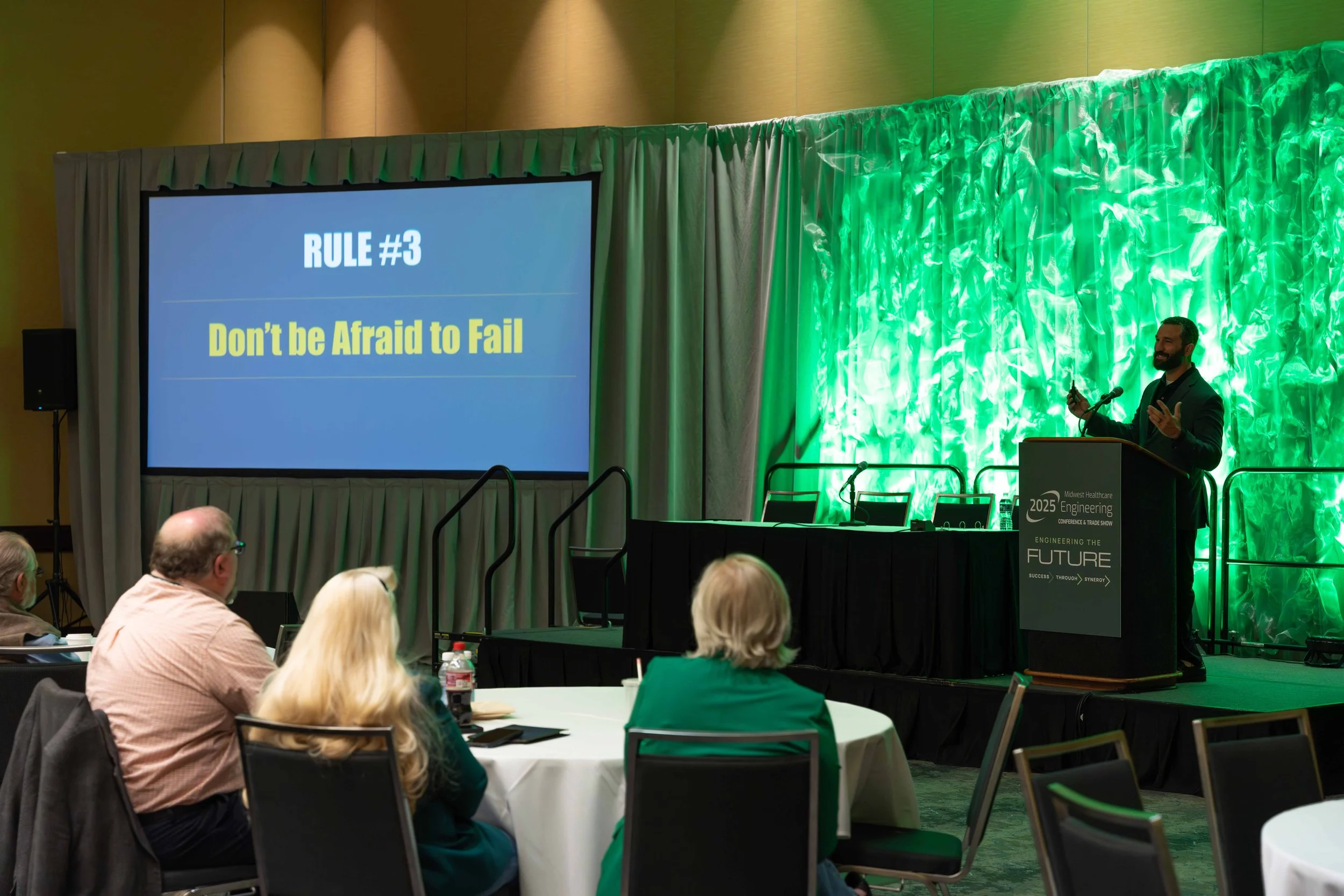 A speaker at a conference podium presenting a slide that reads 'RULE #3 Don't be Afraid to Fail' to an audience seated at round tables in a large conference room with a green illuminated backdrop.
