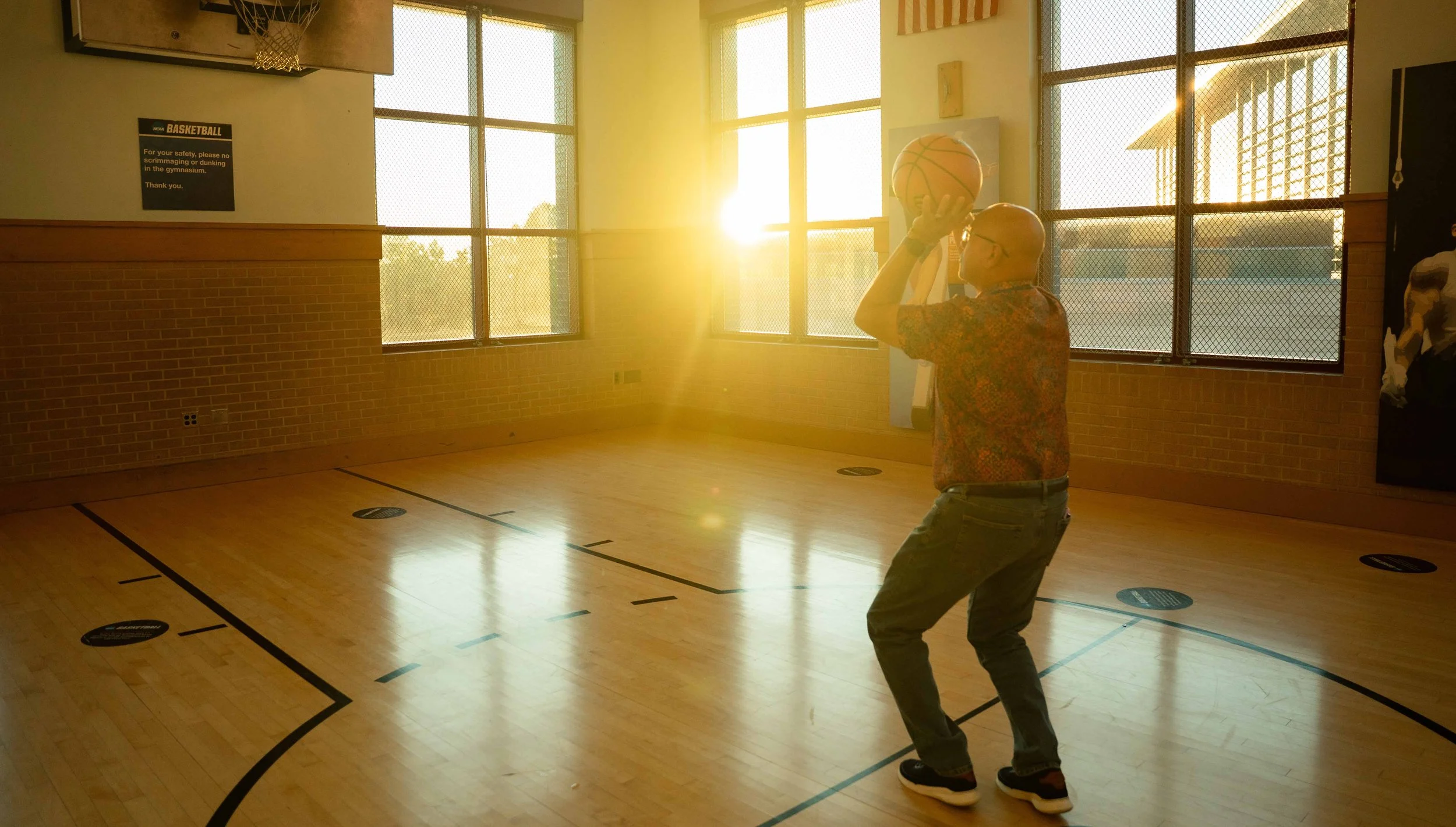 A man wearing glasses and a patterned shirt prepares to shoot a basketball in an indoor gymnasium with large windows and sunlight streaming in.