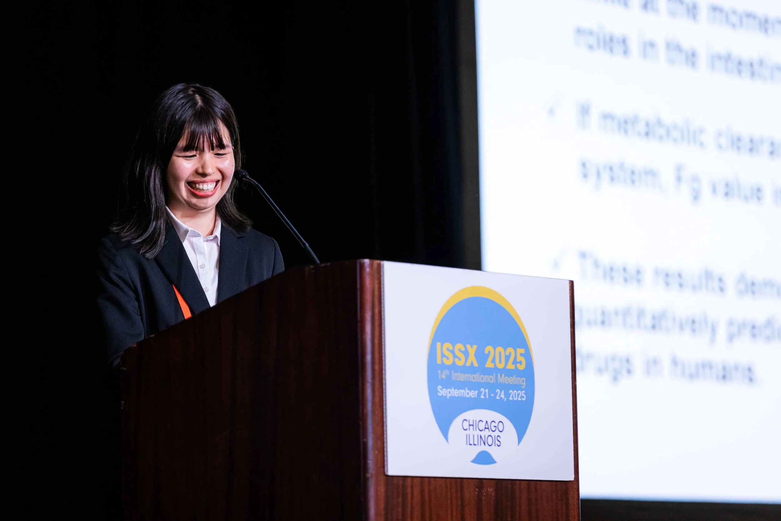 A woman with dark hair, wearing a black blazer, stands at a podium, smiling while giving a speech at the ISSX 2025 conference held in Chicago, Illinois, from September 21 to 24, 2025.