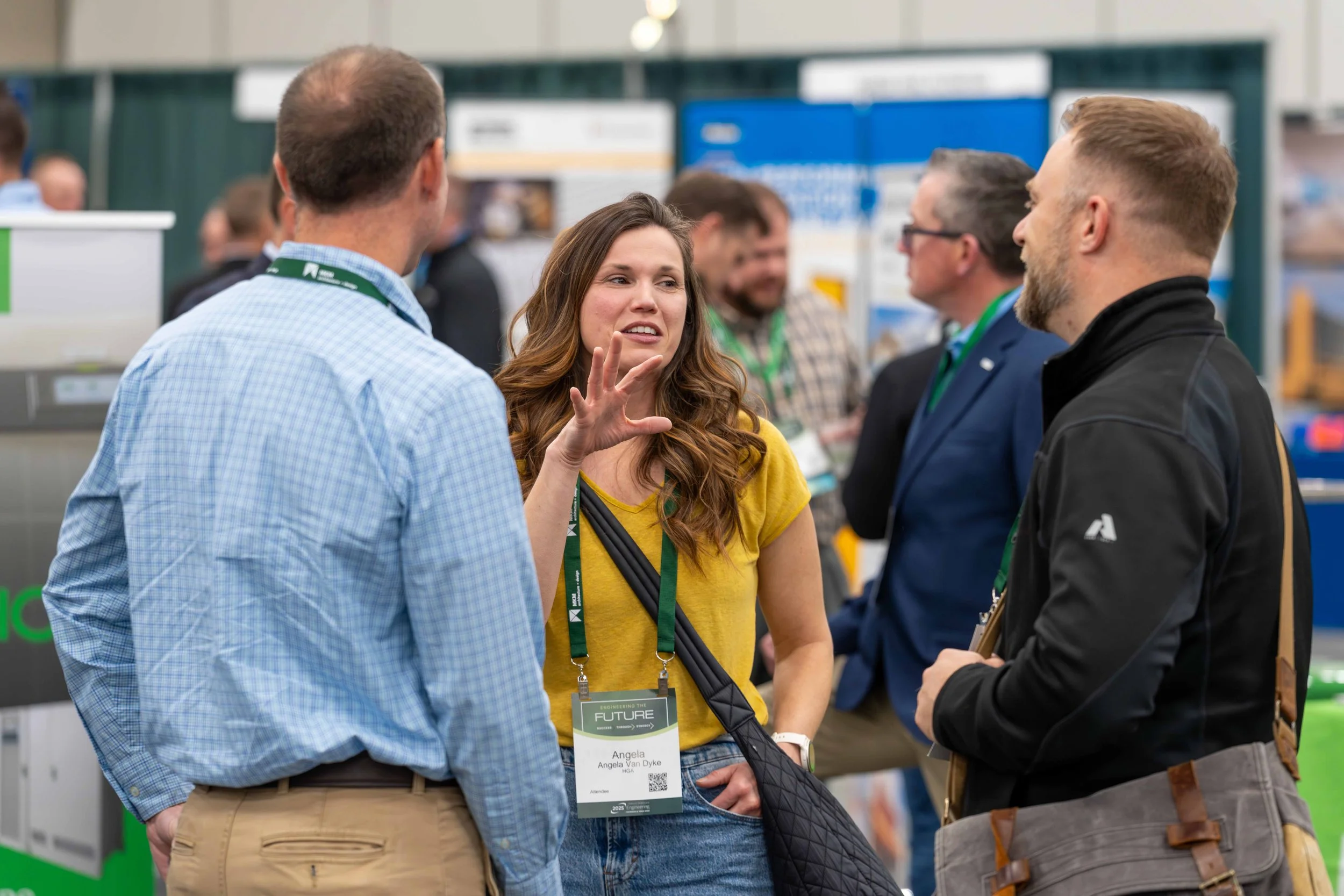 People engaging in conversation at a conference or trade show, with name tags and booths in the background.