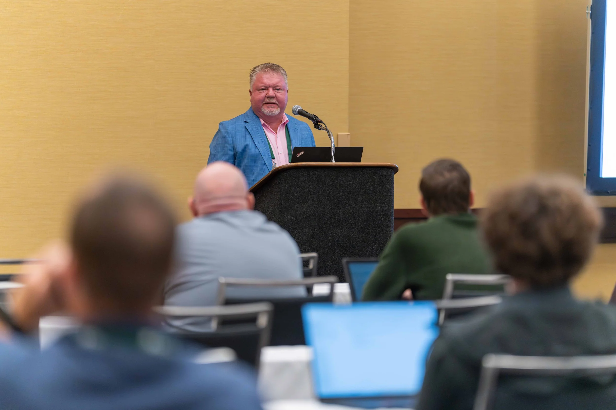A man in a blue blazer speaking at a podium in front of an audience in a conference room.