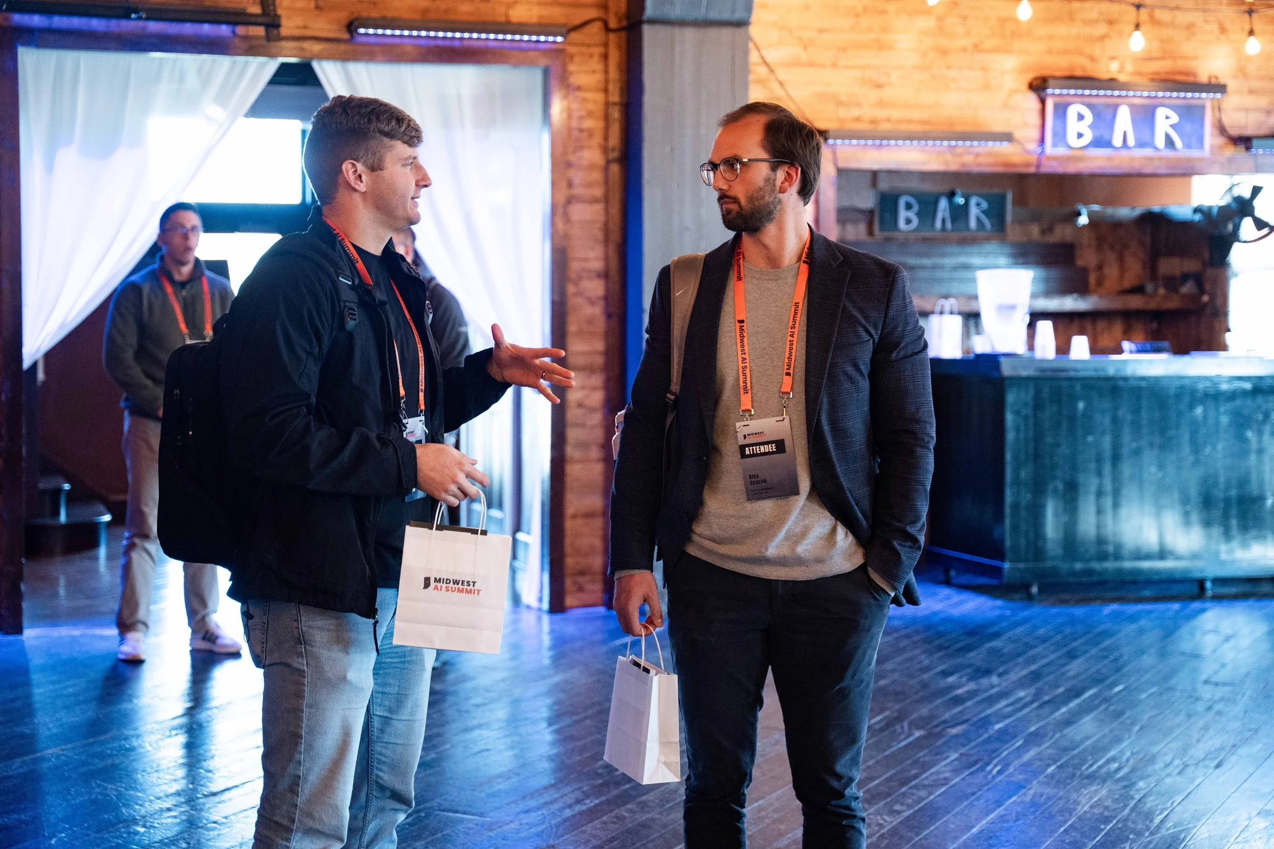 Two men with conference badges and bags are having a conversation at a Midwest AI Summit event in a rustic venue with wood paneling, a bar, and string lights.