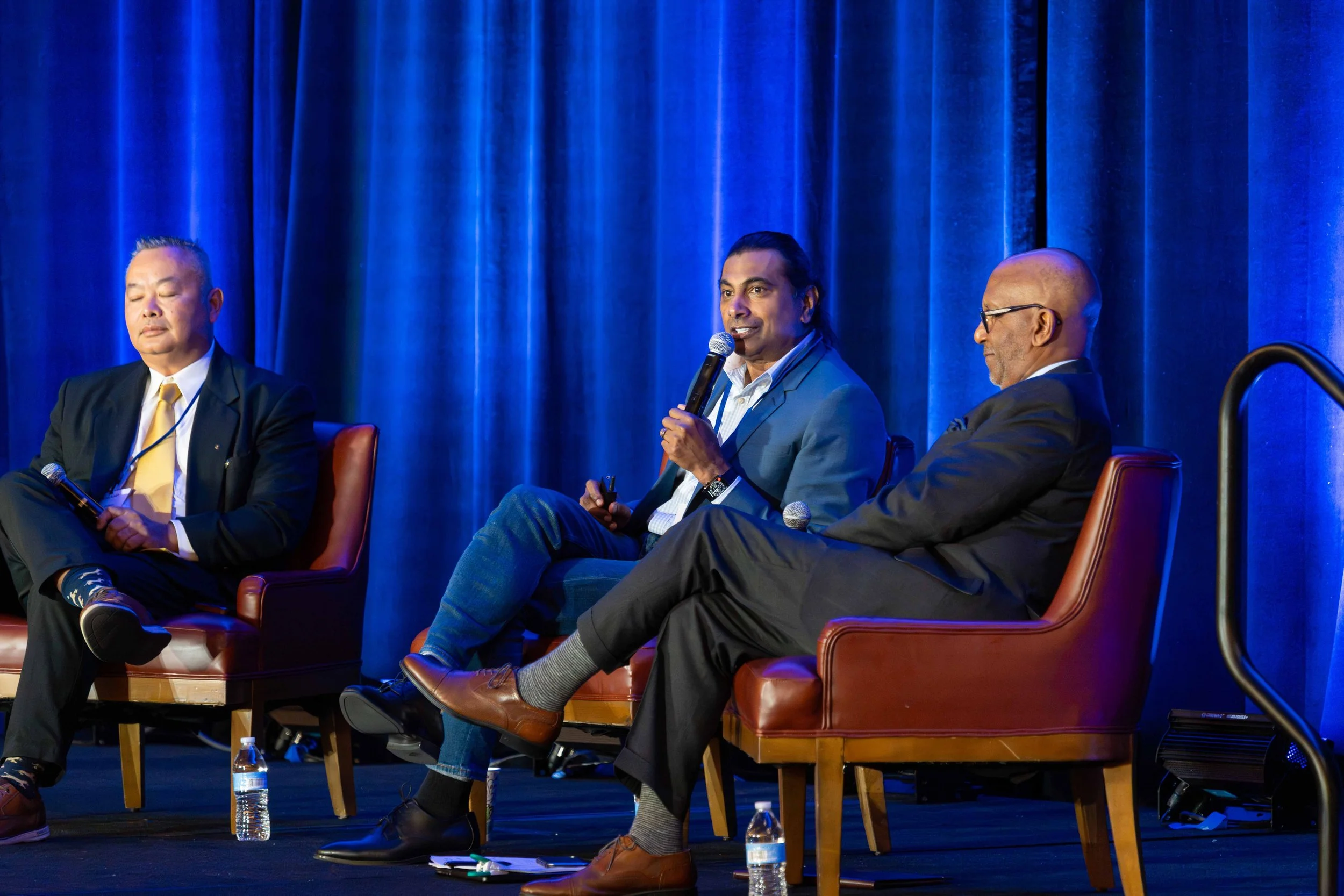 Three men in suits sitting on stage chairs during a panel discussion, with a blue curtain backdrop.