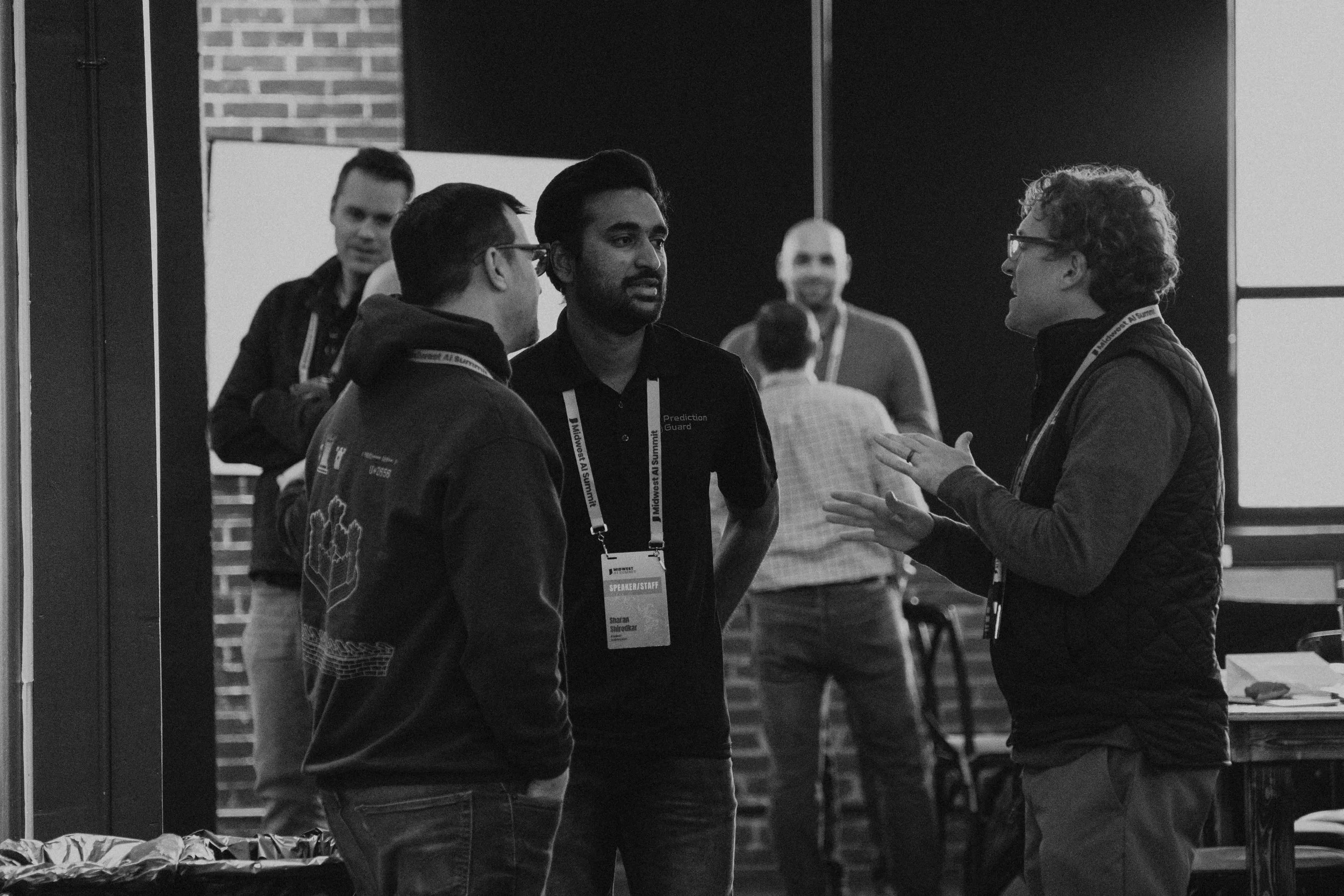 Three men engaged in conversation at a conference, wearing conference badges; other attendees in the background.