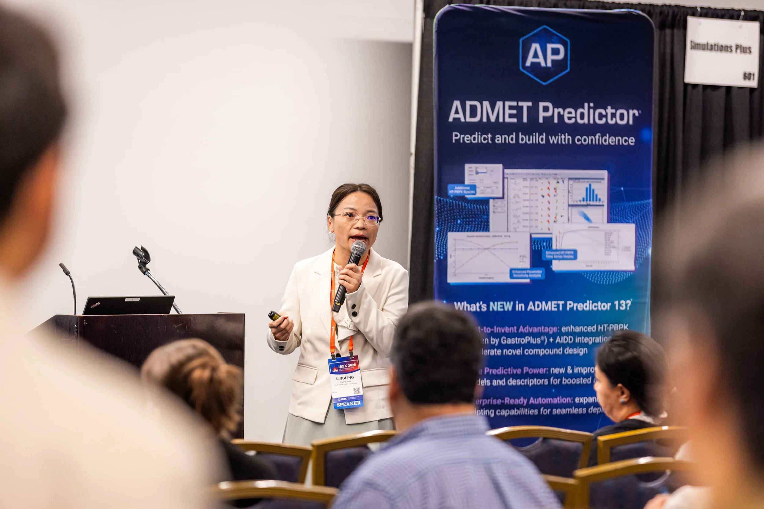 A woman wearing glasses and a white blazer is giving a presentation at a conference. She is holding a microphone and a remote control, standing in front of a blue banner that reads 'ADMET Predictor' with graphical data displays and text about new fea