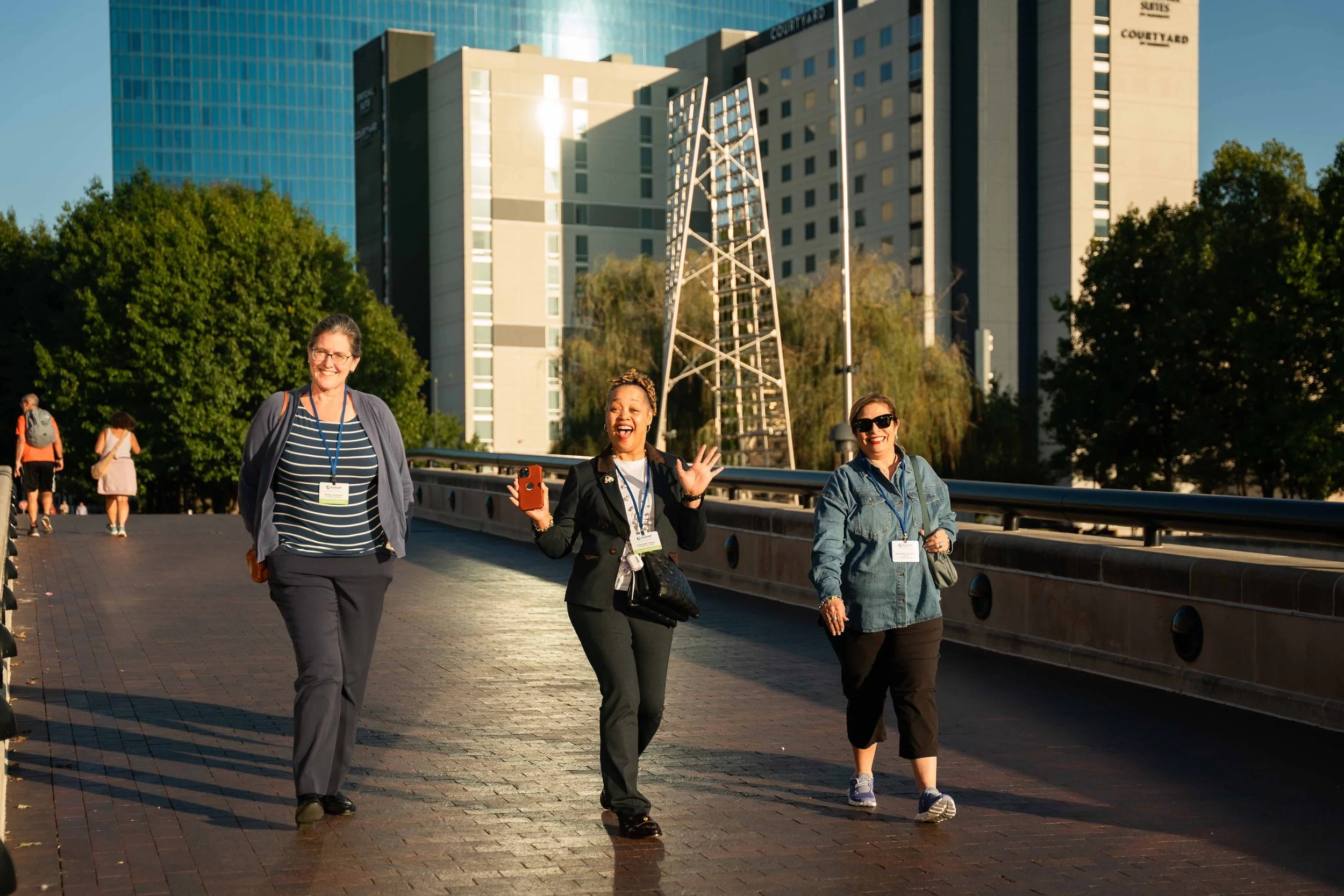 Three women walking outdoors in an urban area, smiling and talking, with tall buildings and trees in the background, during the daytime.